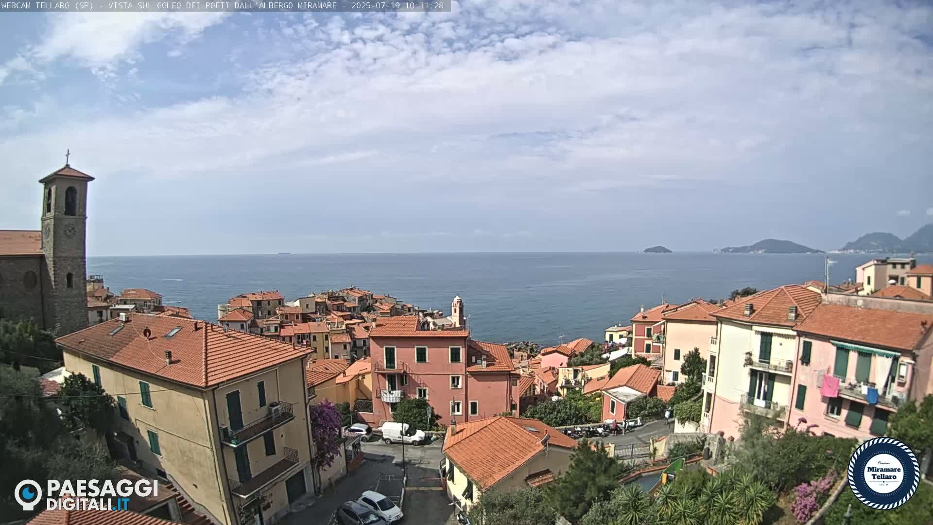 A coastal village with red-tiled roofs overlooks a calm sea under a partly cloudy sky.