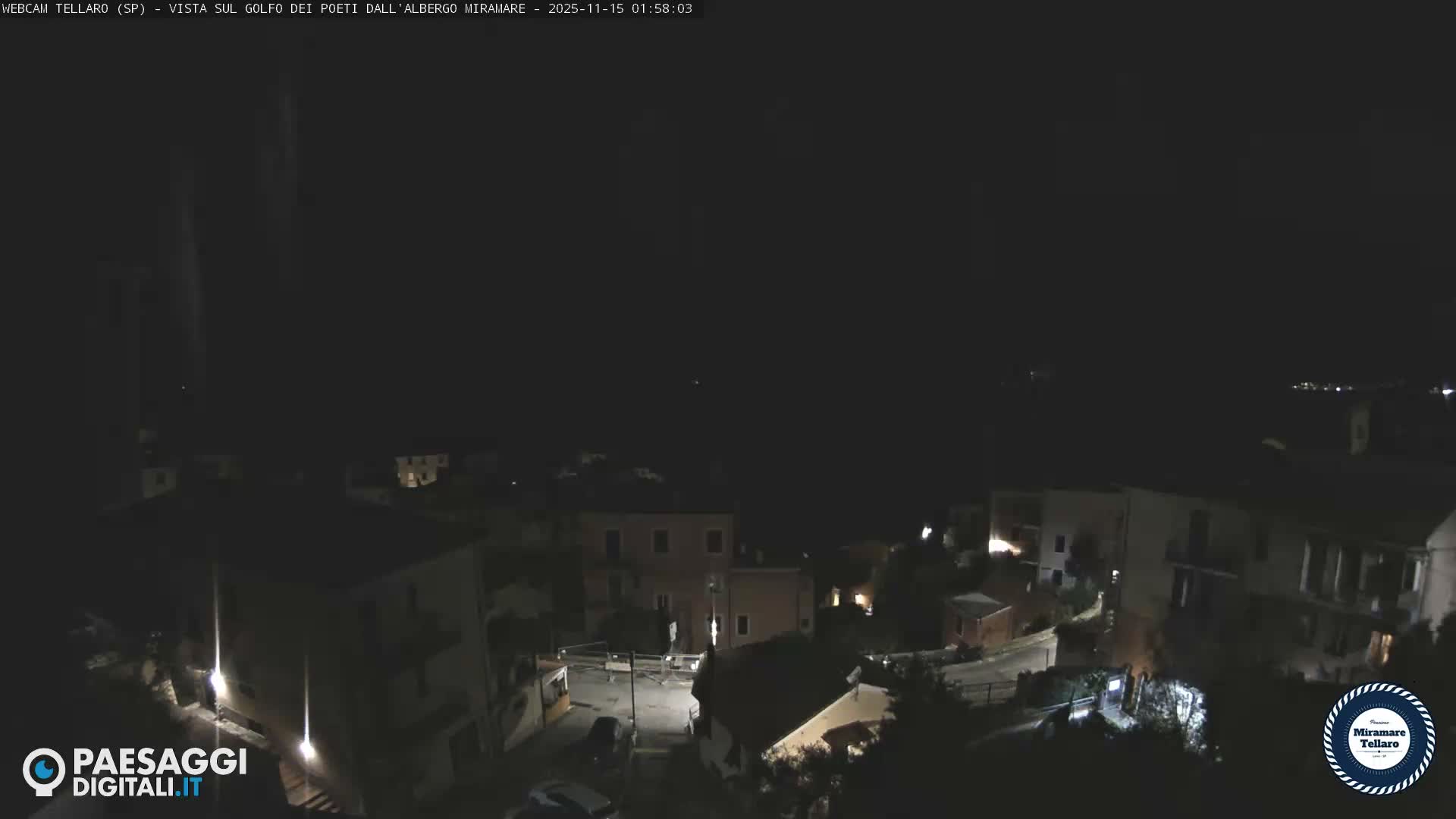 The image shows a nighttime view of a town with illuminated buildings and streetlights, overlooking a body of water with distant lights, under a clear sky.