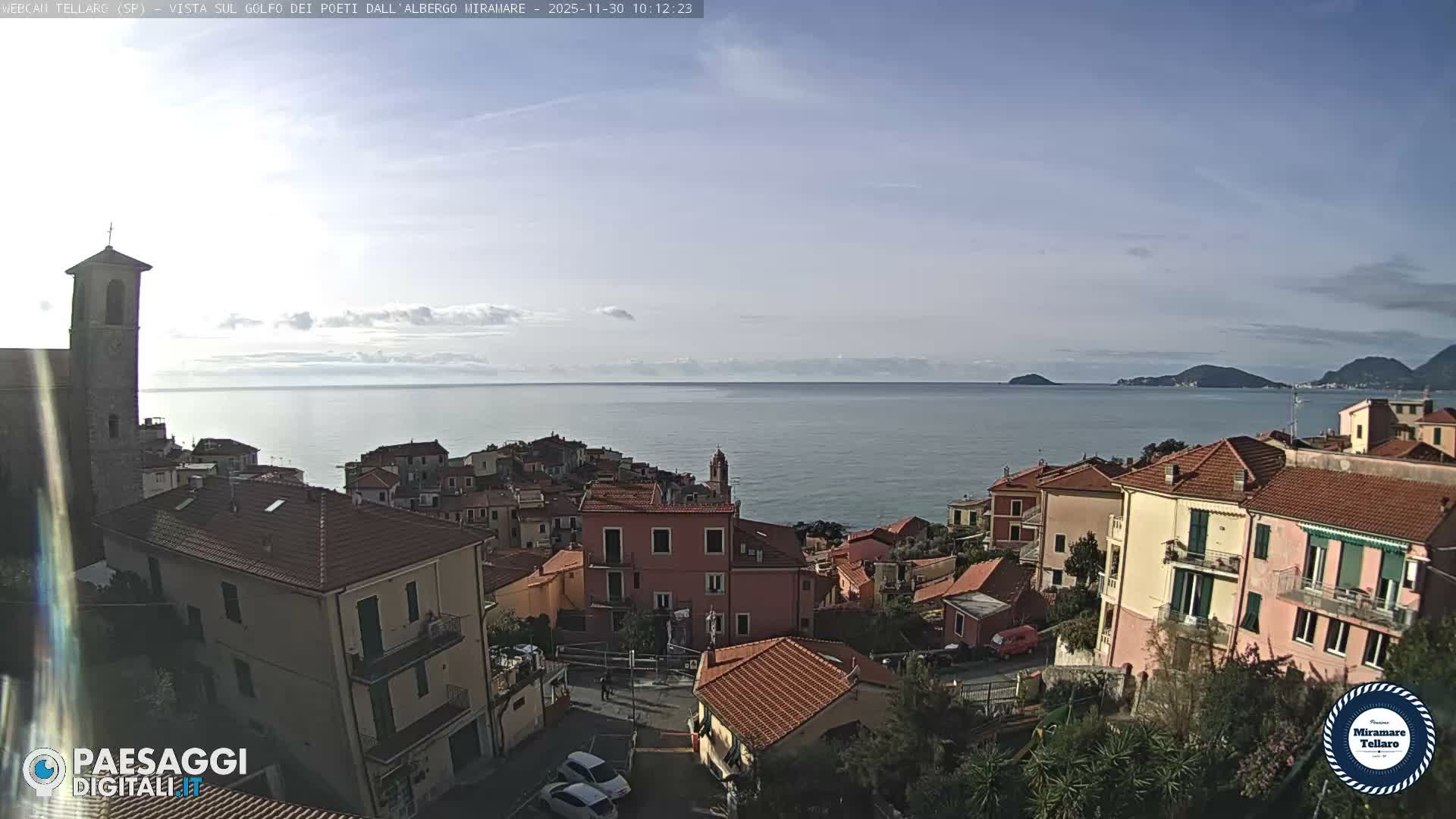 A charming coastal town with colorful buildings and a prominent bell tower cascades down a hillside towards a calm bay featuring distant islands and hills, all under a partly cloudy sky.