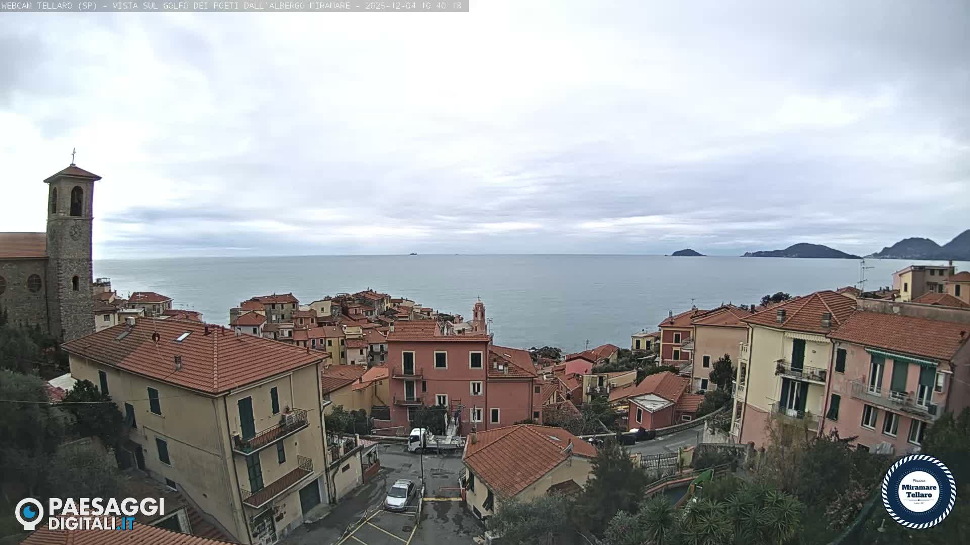 An overcast day reveals a picturesque Italian coastal village with terracotta-roofed houses and a prominent stone church tower cascading down to a calm sea, with distant islands and mountains visible on the horizon.