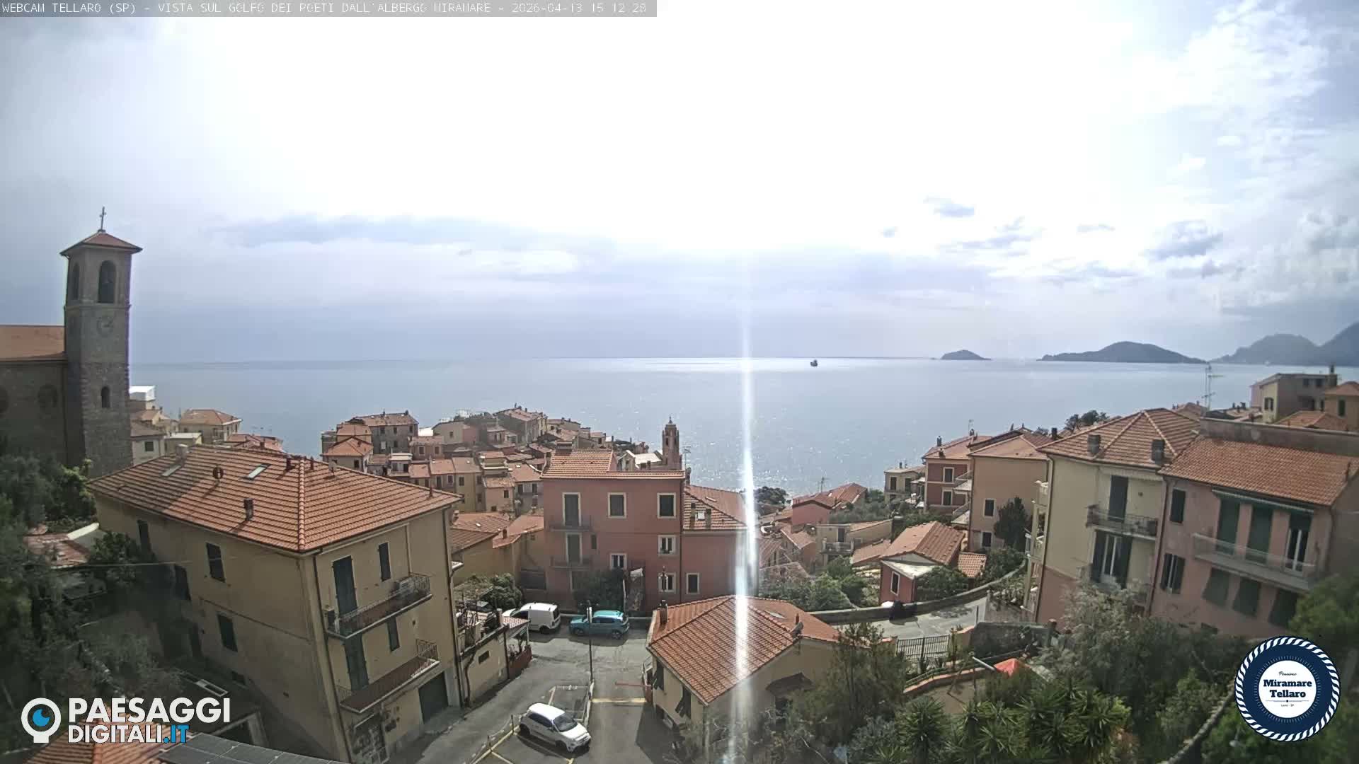 An overcast day reveals a picturesque Italian coastal village with terracotta-roofed houses and a prominent stone church tower cascading down to a calm sea, with distant islands and mountains visible on the horizon.