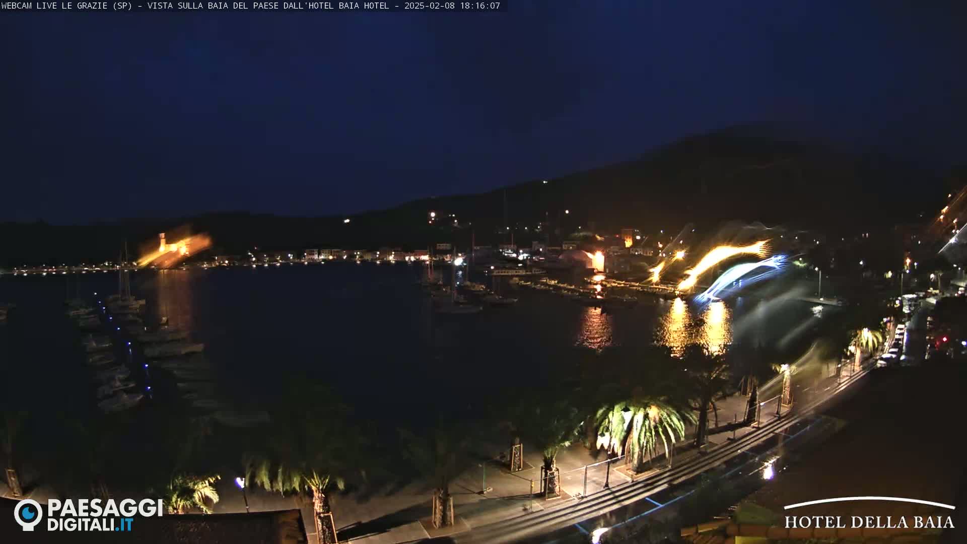 A nighttime view of a harbor with boats, illuminated buildings on a hillside, and a fireworks display over the water.