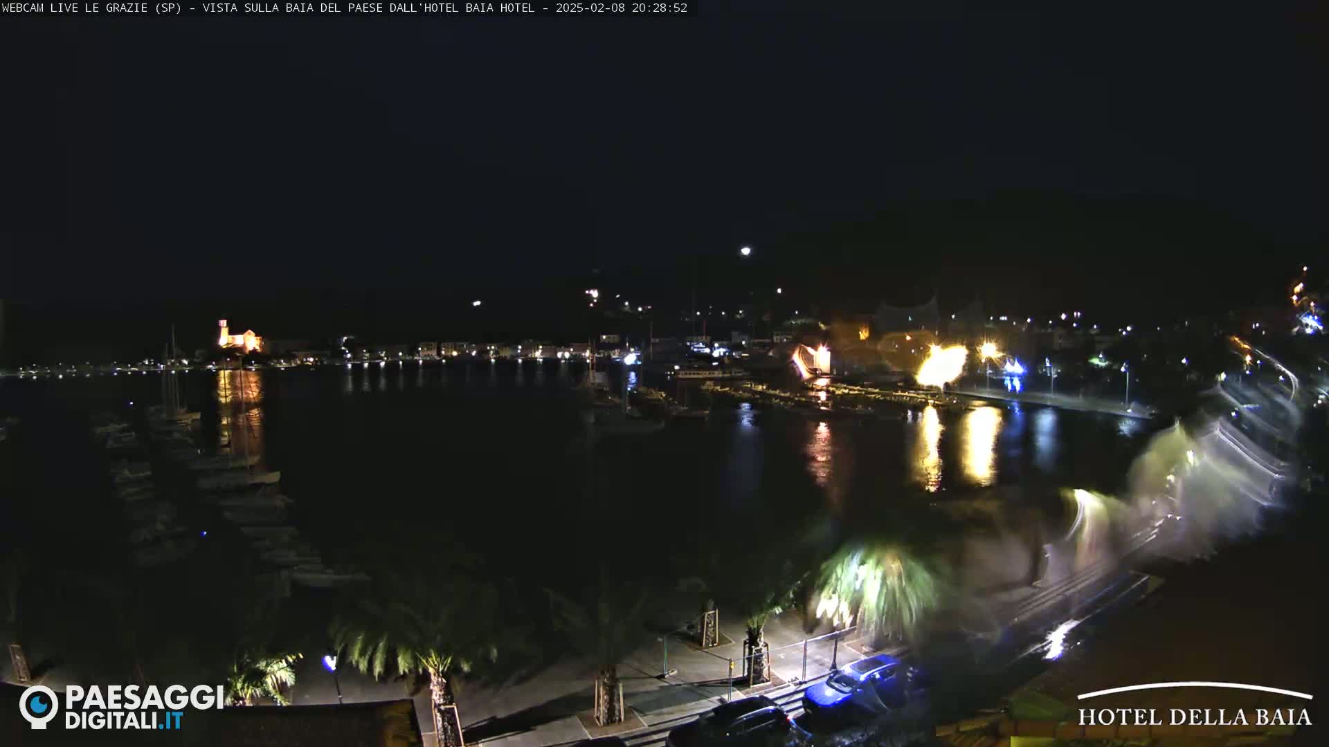 A nighttime harbor scene shows a town illuminated along a shoreline with boats in the water; fireworks are exploding over the water.