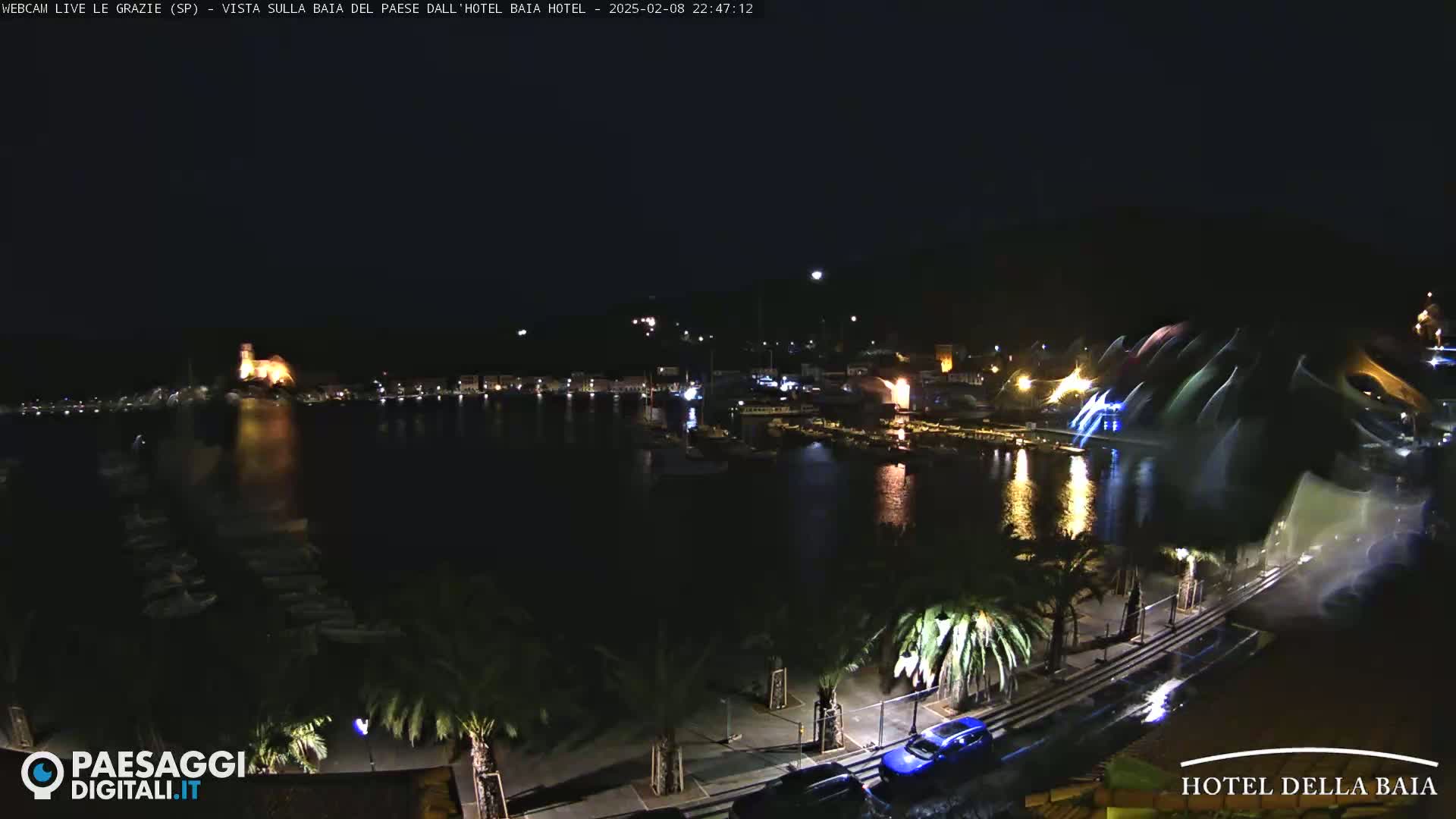 A nighttime harbor scene shows a waterfront town illuminated with lights, palm trees lining the streets, and a water feature spraying colorful light into the air.