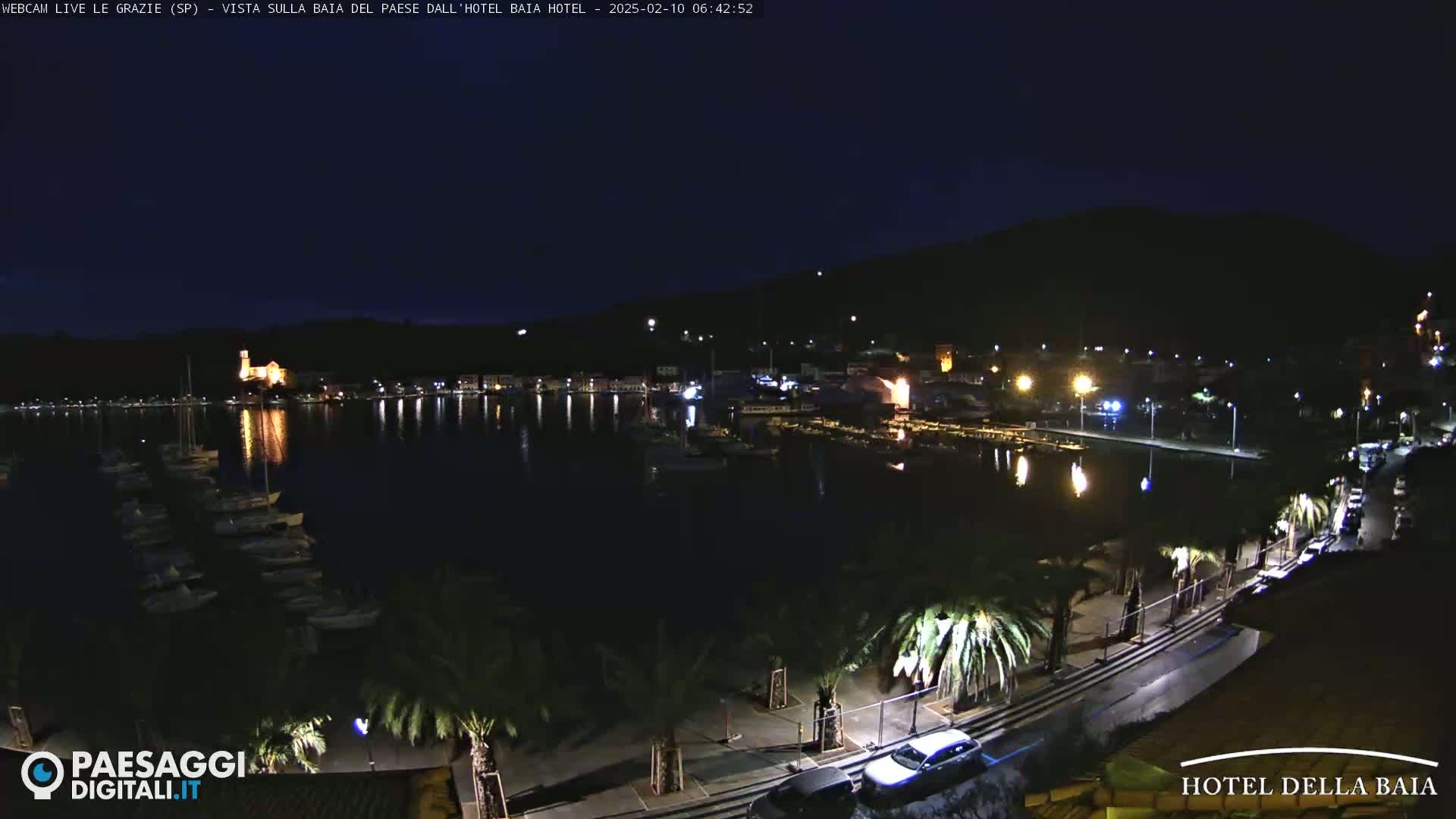 A nighttime view of a harbor, with boats moored and town lights reflecting on the water, under a dark, clear sky.