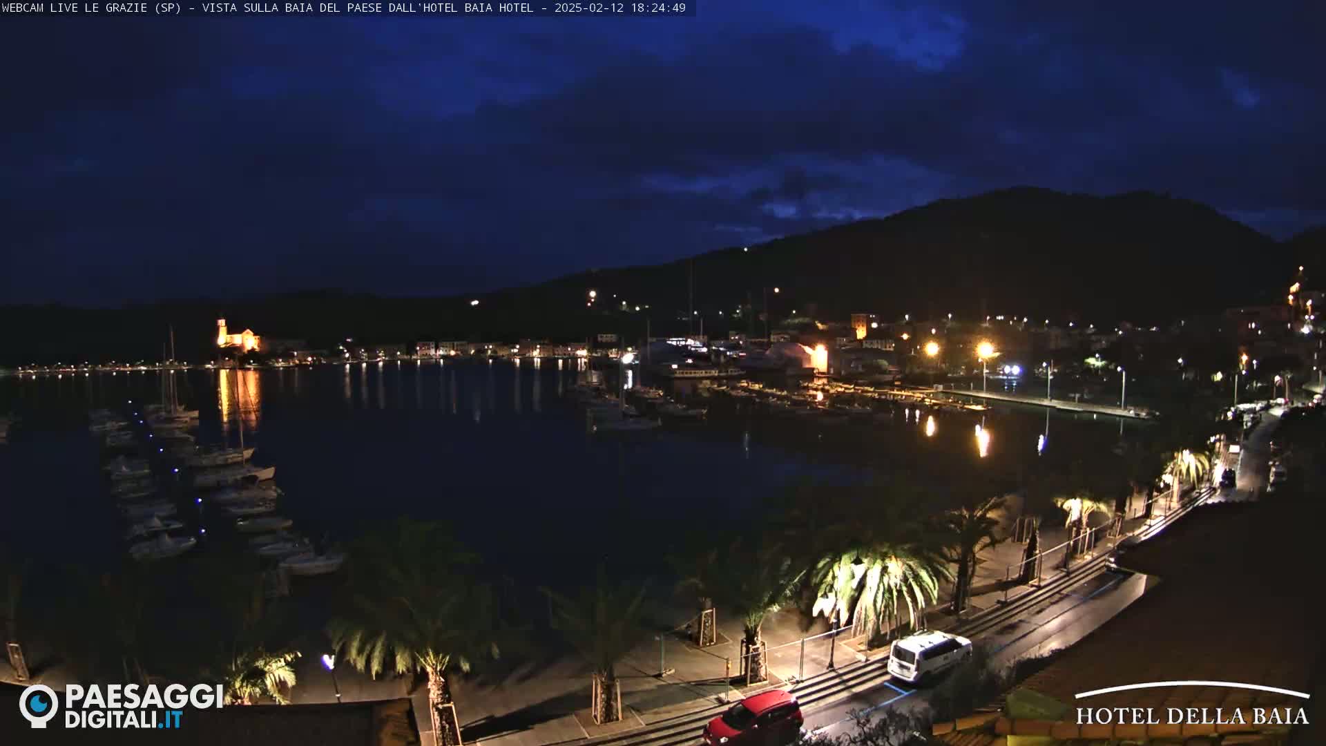 A nighttime harbor scene shows a town illuminated along a calm body of water, where boats are docked, under a dark, slightly cloudy sky.