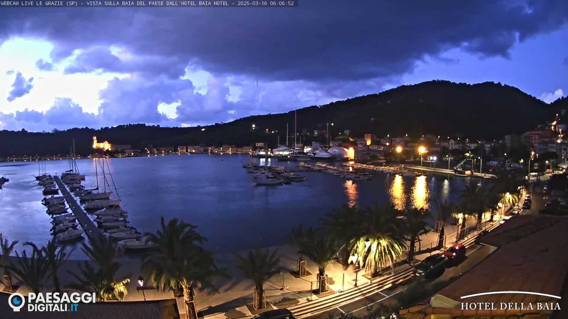 A nighttime view of a harbor filled with boats, lined with palm trees and buildings along a waterfront promenade under a partly cloudy sky.