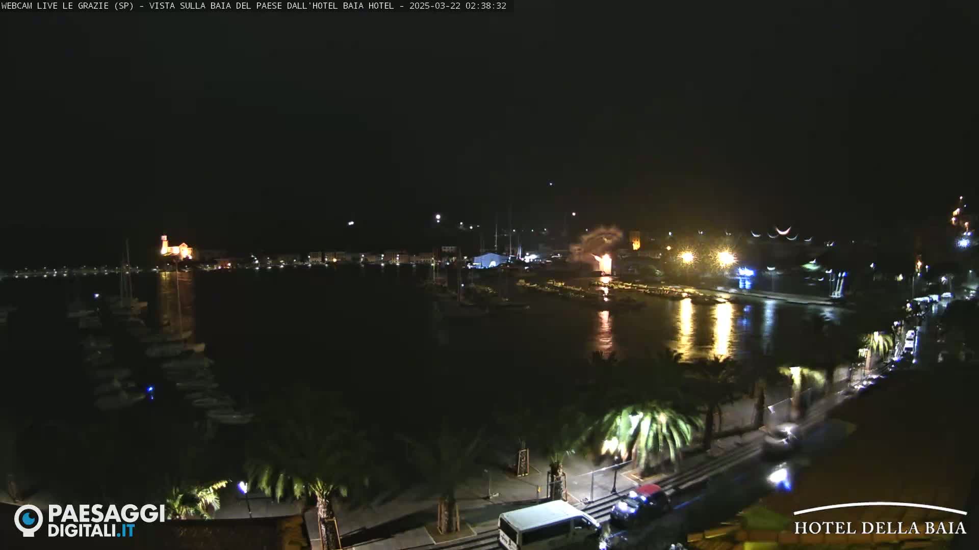 A nighttime harbor scene with fireworks exploding over the water, illuminated buildings, and palm trees lining a roadway.