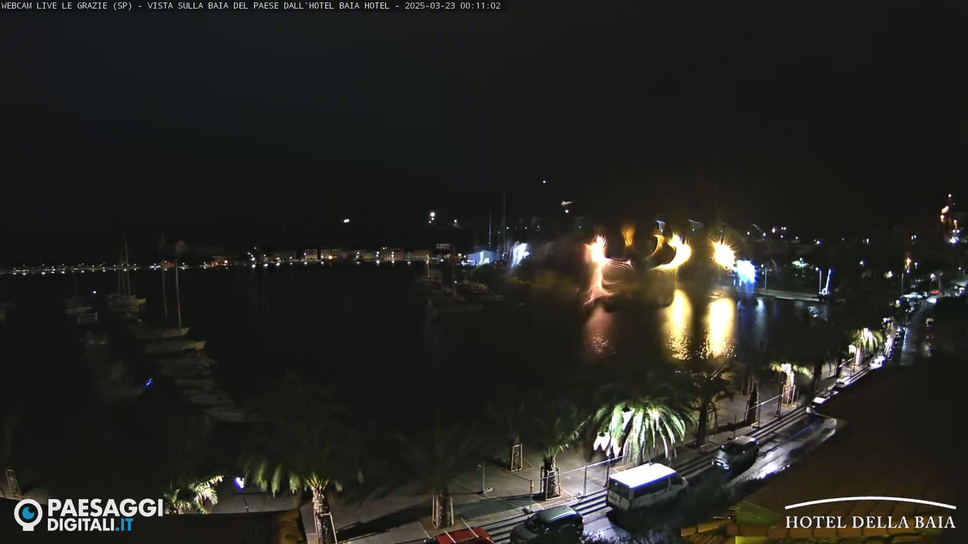 A nighttime harbor scene with fireworks exploding over the water, palm trees lining a roadway, and boats moored at the docks.