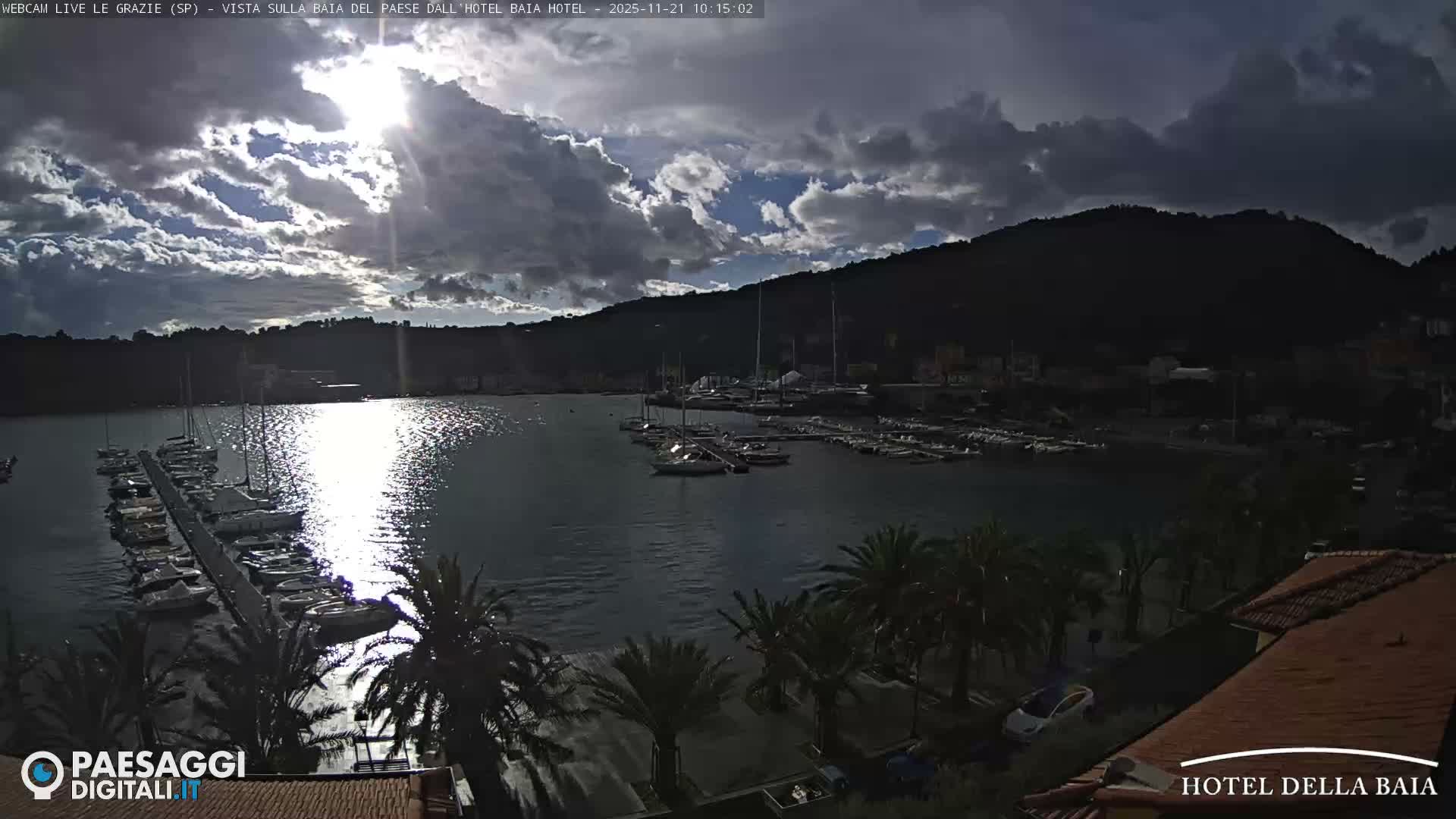 A scenic bay features numerous boats docked along piers, with palm trees and distant hills under a partly cloudy sky where bright sunlight reflects intensely off the water.