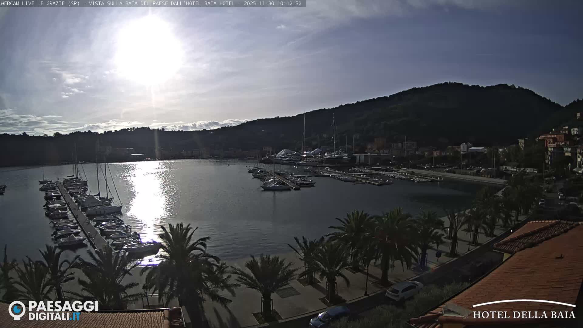A bright, sunny day with scattered clouds illuminates a picturesque coastal bay filled with numerous docked boats, a bustling waterfront town, lush green hills, and palm trees along the shore.