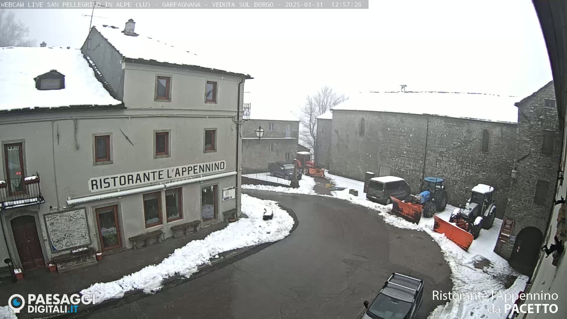 A snow-covered village square with several buildings, parked cars, and snow-clearing equipment under a light snowfall.