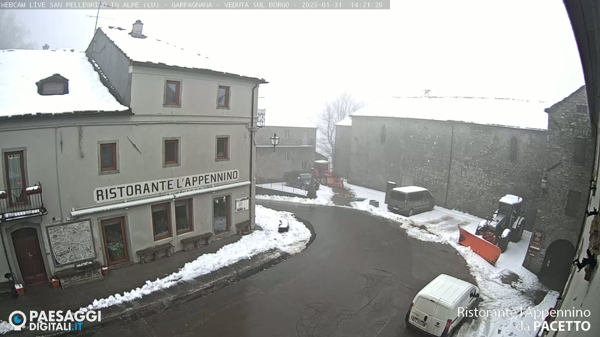 A snow-covered village square with several buildings, a snow plow, and parked vehicles in foggy conditions.