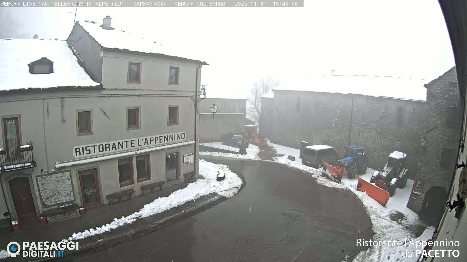 A snow-covered village square with several buildings, some parked vehicles, and snow removal equipment in foggy conditions.