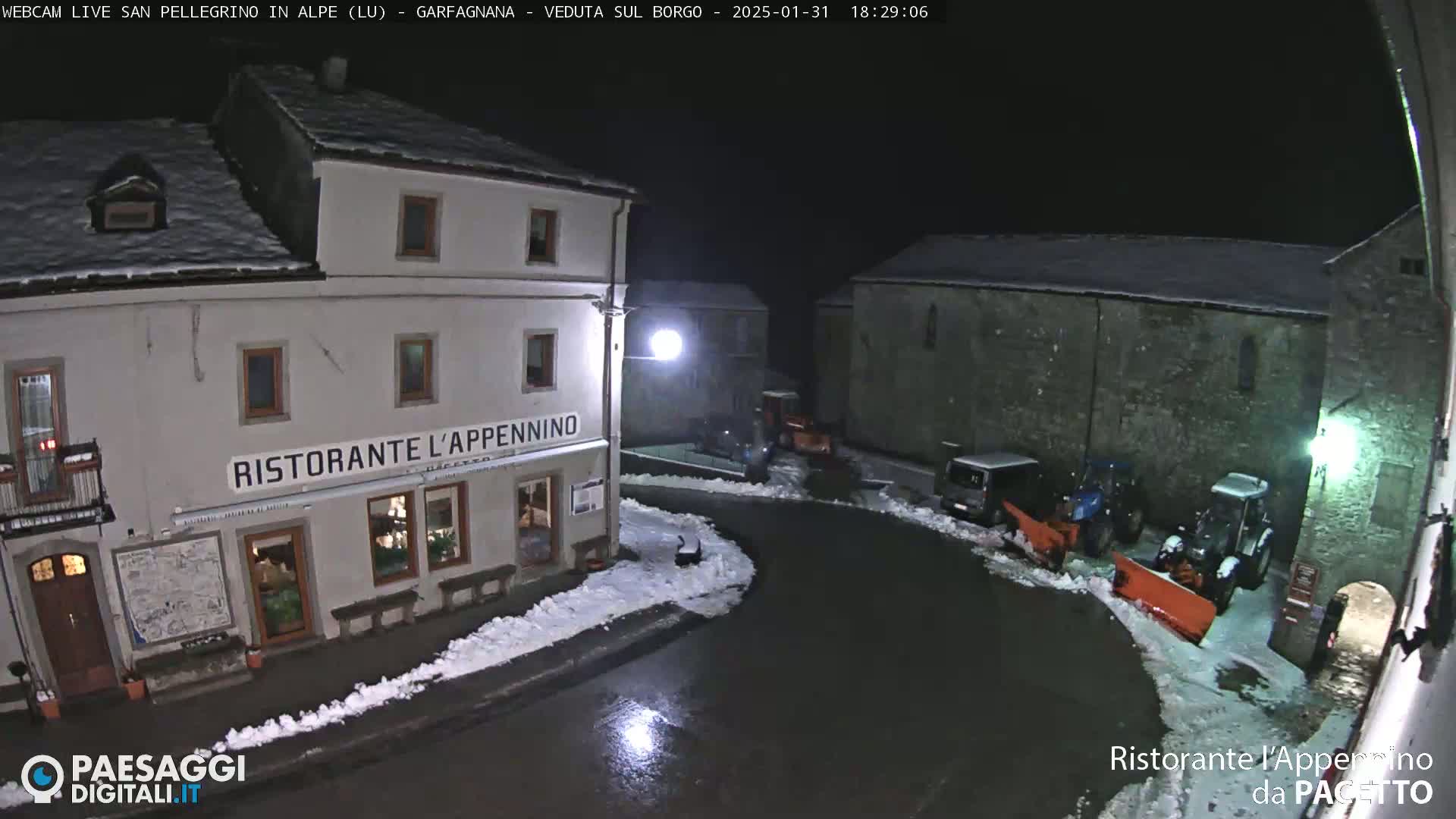 A snow-covered village square at night, illuminated by streetlights, features a restaurant and several snowplows clearing wet roads.