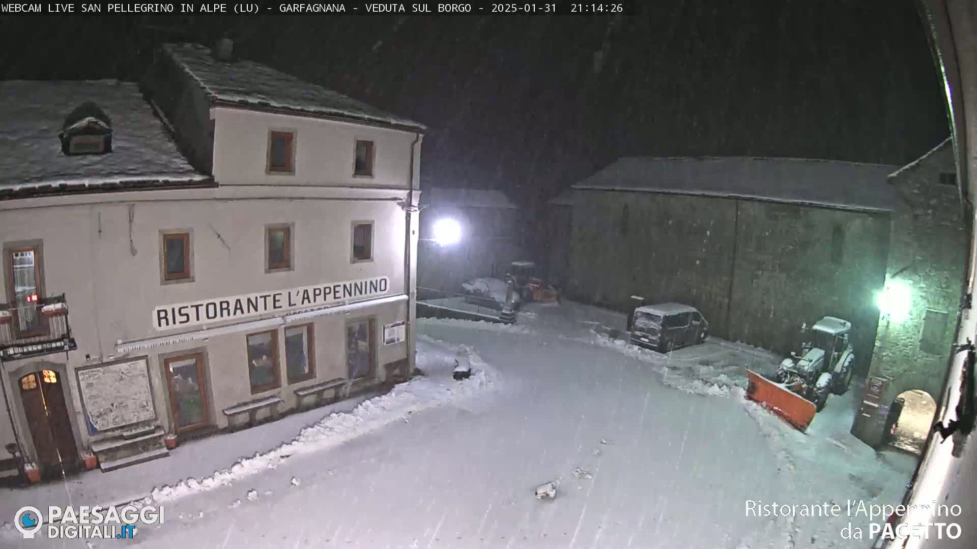 A snow-covered town square at night, featuring a restaurant, parked vehicles, and snow removal equipment under falling snow.