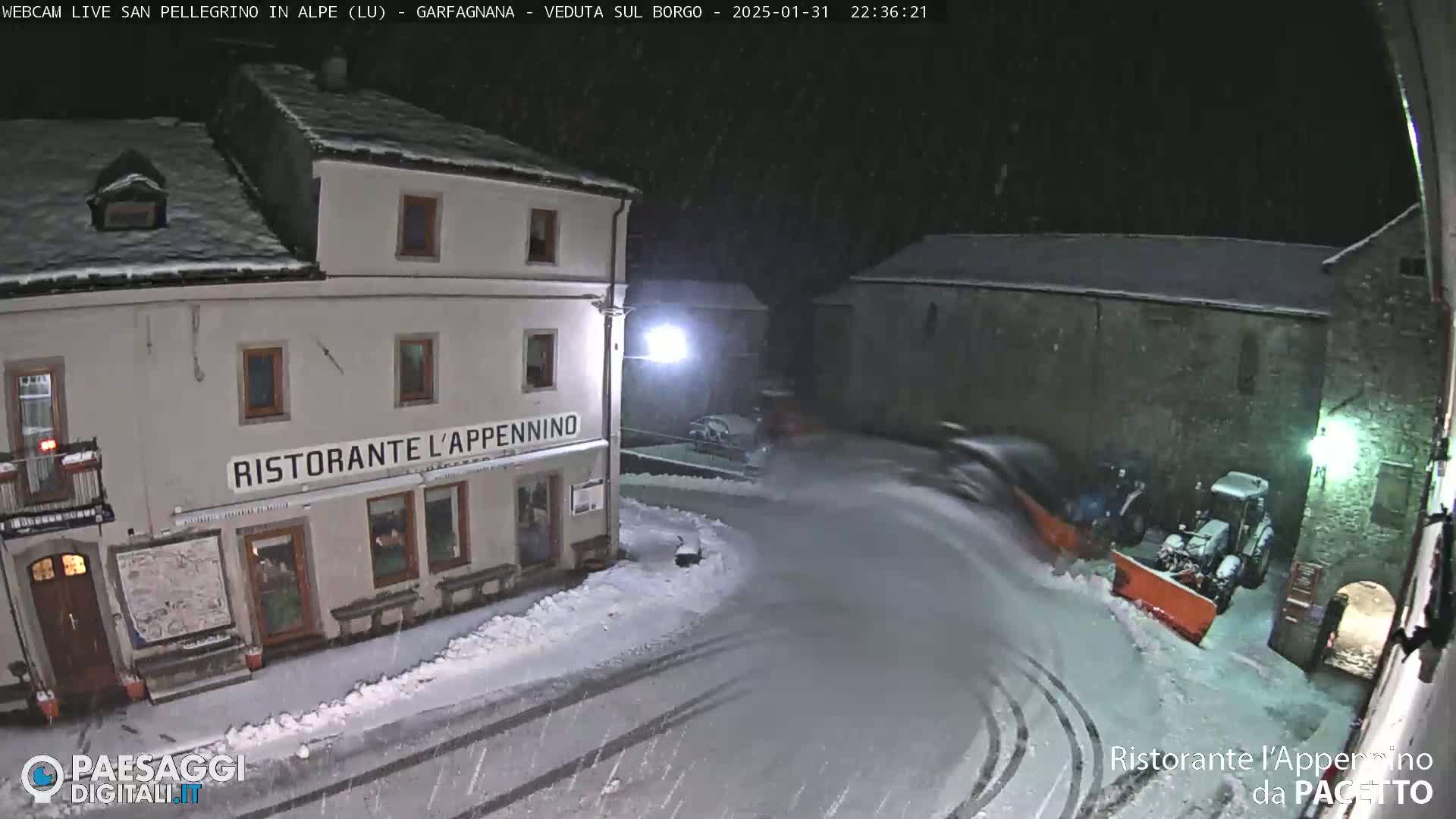 A snow-covered town square at night, with a snowplow clearing a curved road in front of a restaurant.