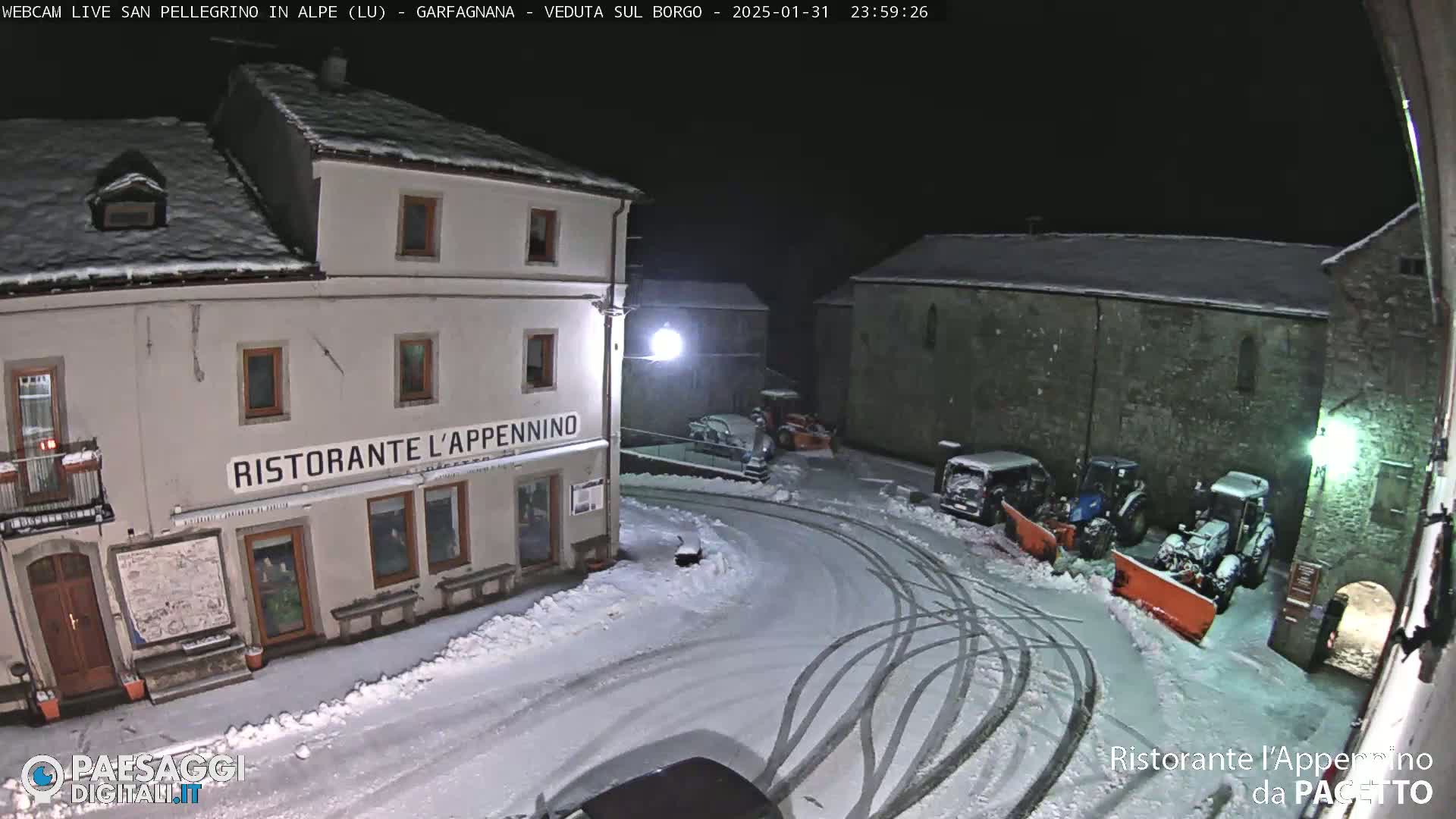 A snow-covered town square at night, with several vehicles, including snowplows, parked near buildings.