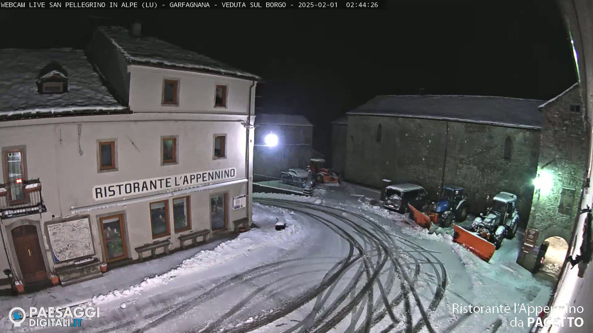 A snow-covered town square at night, with several vehicles and snow-clearing equipment parked near buildings.