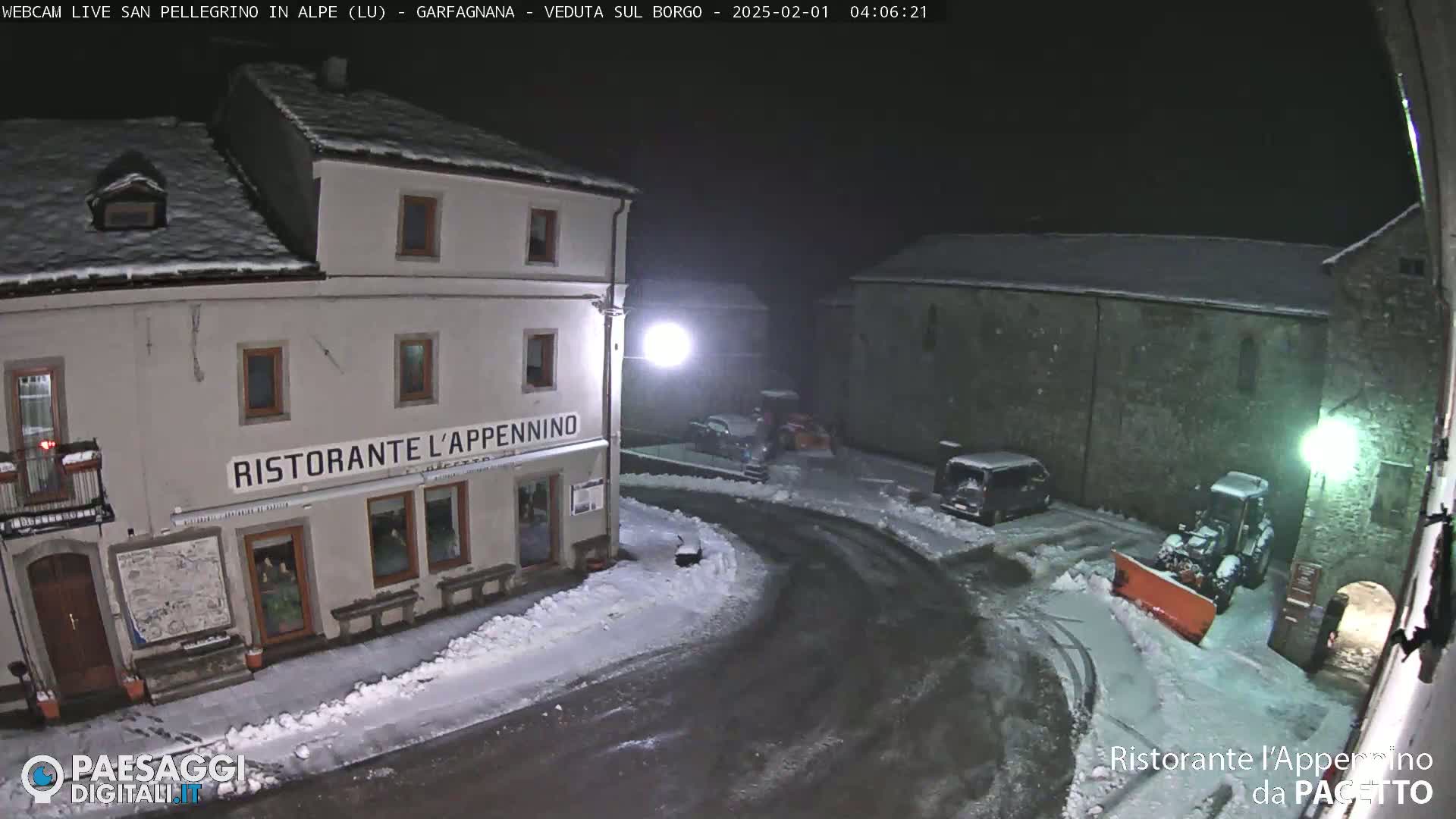 A snow-covered town square at night, with a restaurant and several parked vehicles, is being plowed by a snowplow.