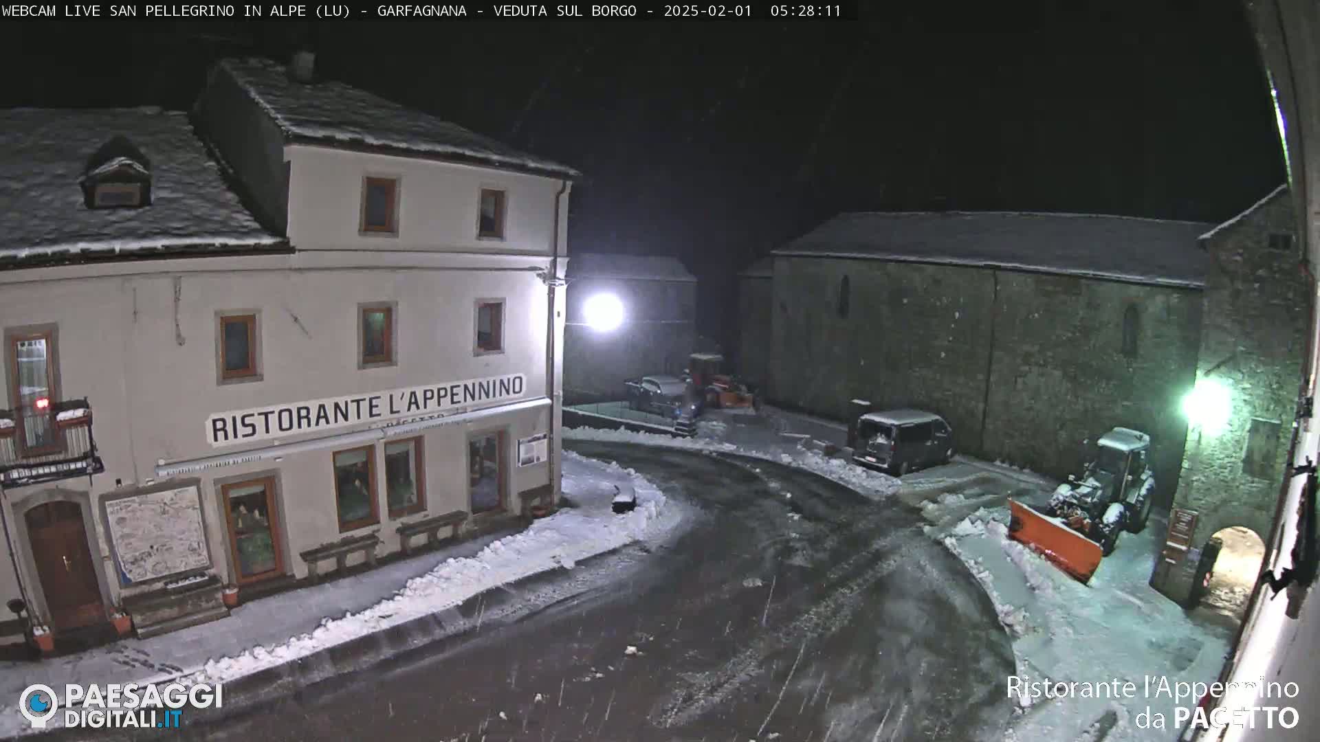 A snow-covered town square at night, showing several buildings, parked cars, and a snowplow.