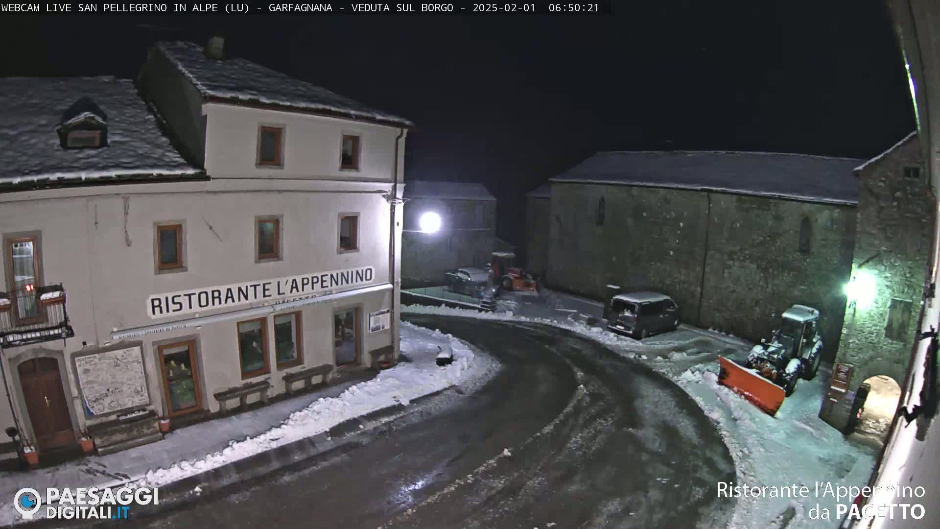 A snow-covered town square at night, with a restaurant, parked vehicles, and a snowplow.
