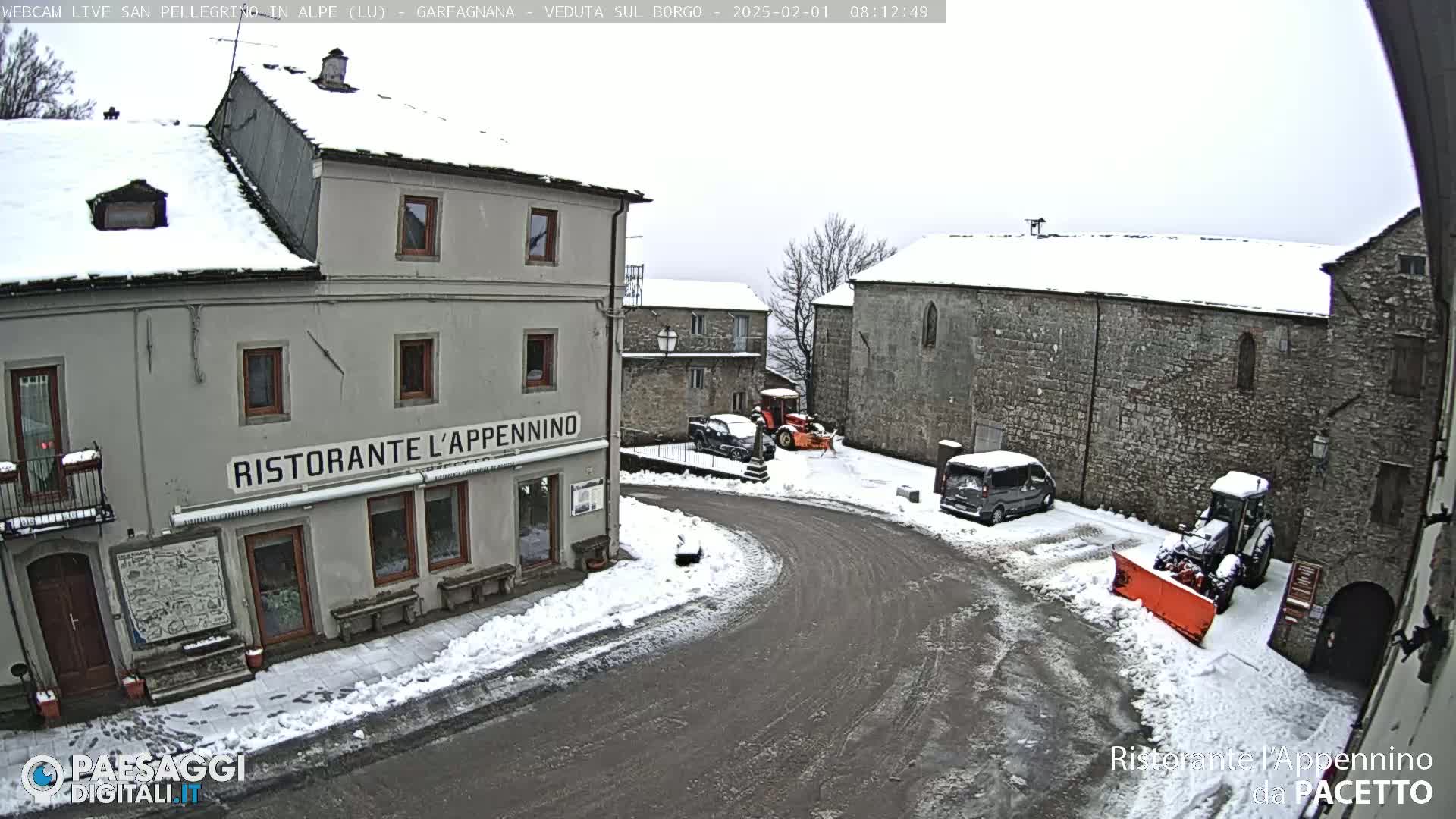 A snow-covered village square with several buildings, vehicles, and snow-removal equipment.