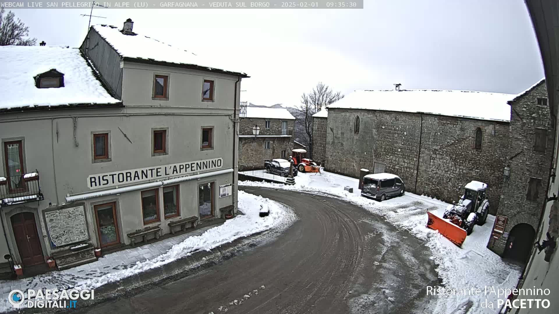 A snow-covered village square with several buildings, vehicles, and snow removal equipment on an overcast day.