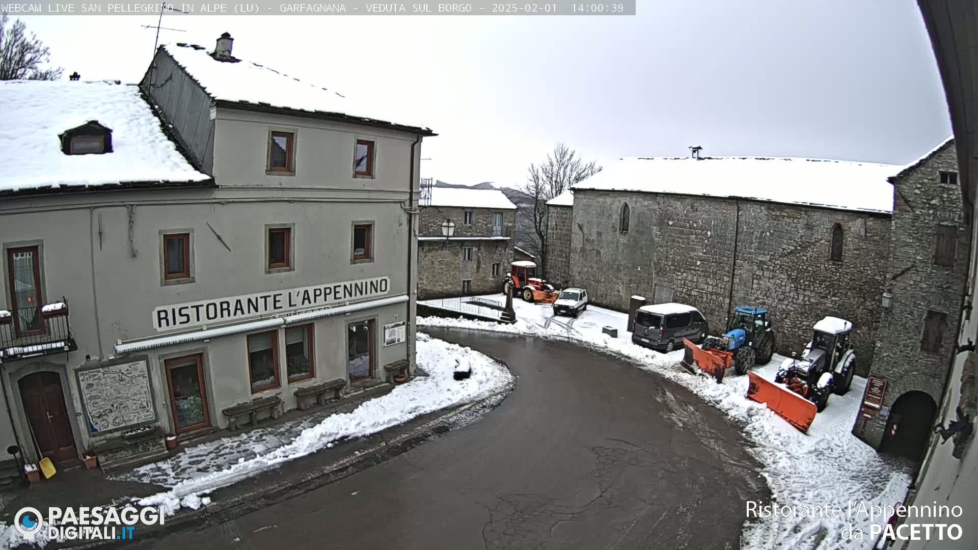 A snow-covered village square with several buildings, some snow-clearing vehicles, and a few parked cars under an overcast sky.