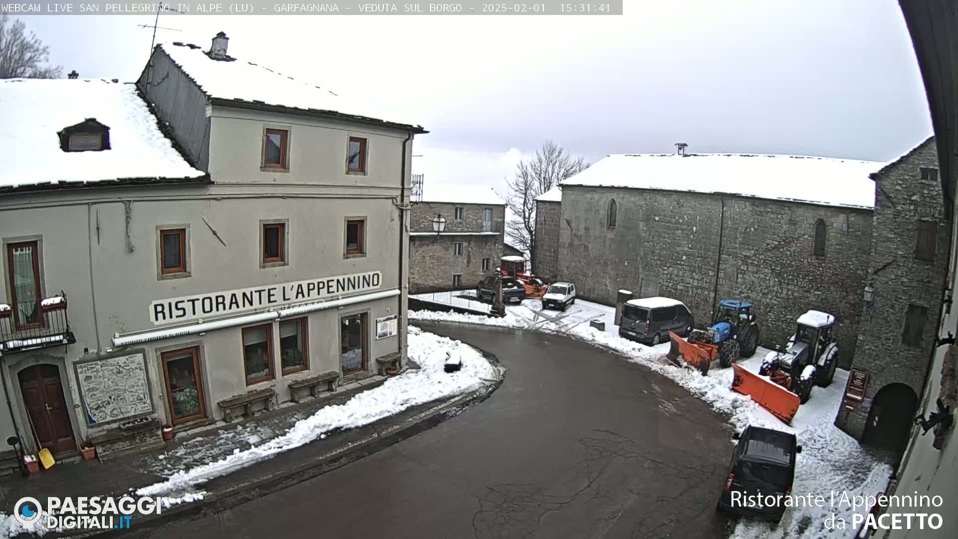 A snow-covered village square with several buildings, parked cars, and snow removal equipment under an overcast sky.