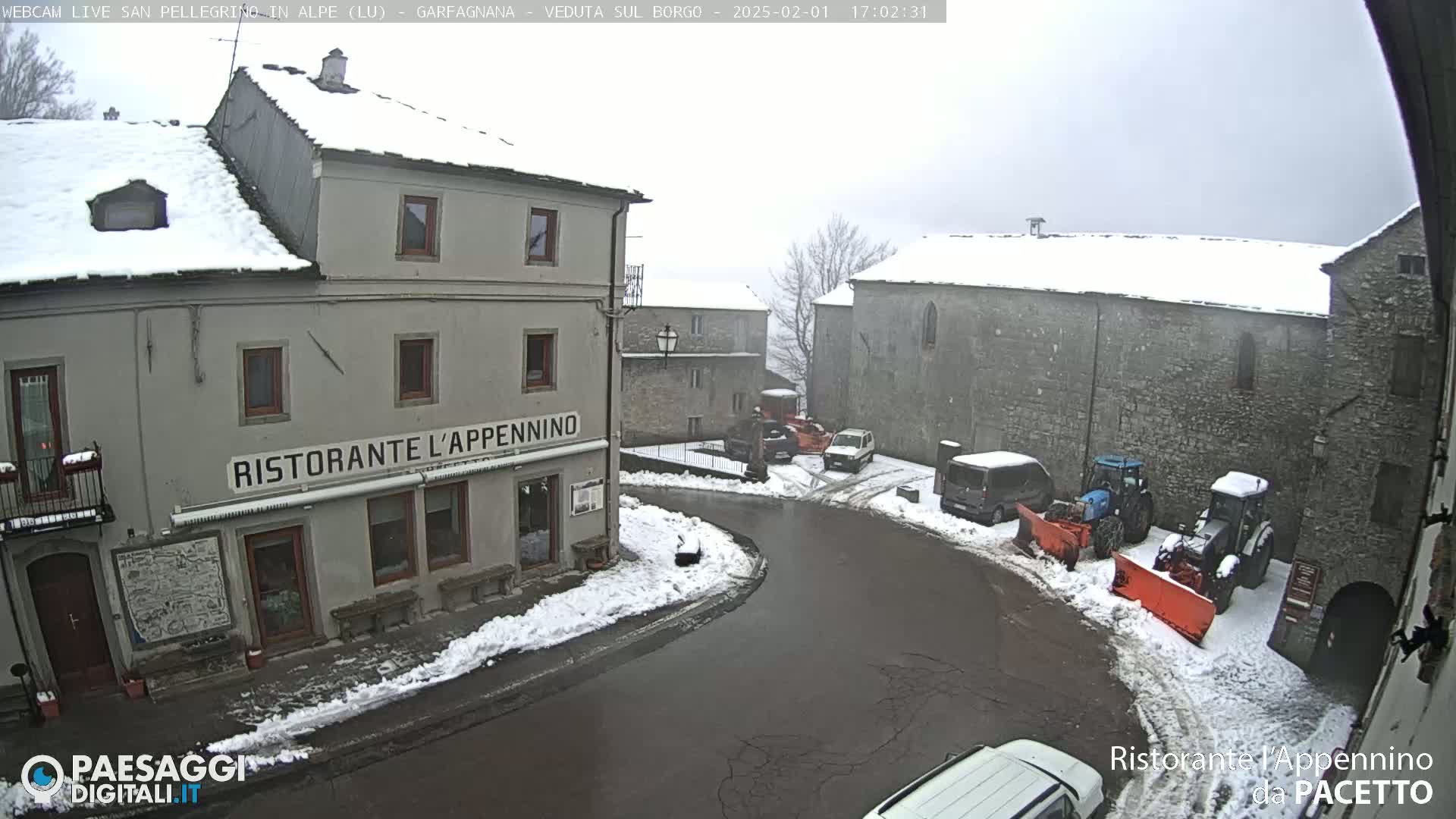 A snow-covered town square with several vehicles and snow-removal equipment, viewed from an elevated position on a cloudy day.