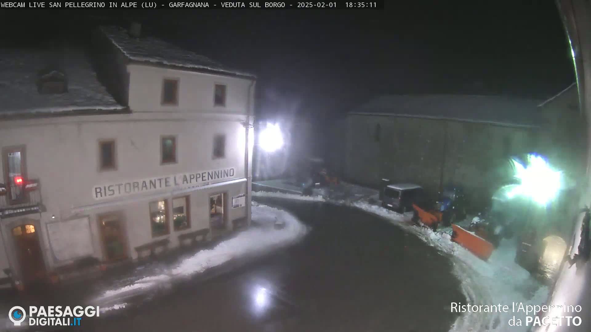 A snow-covered town square at night, with a snowplow clearing the road in front of a restaurant.
