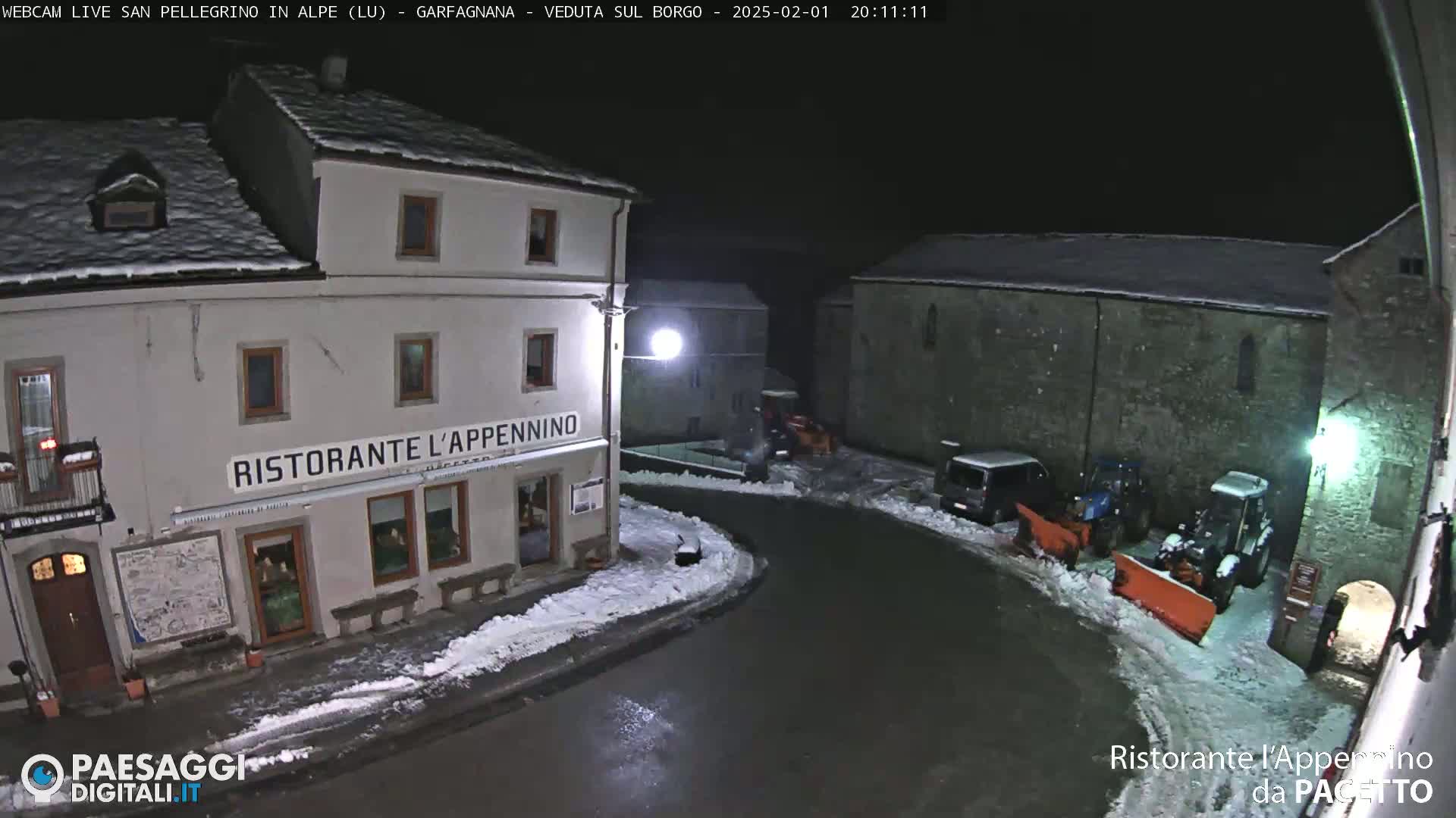 A snow-covered village square at night, with a restaurant and snowplows visible.