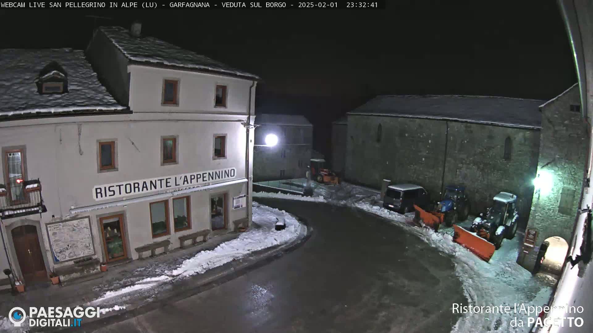 A snow-covered village square at night, illuminated by streetlights, shows several buildings, parked vehicles, and snowplows.