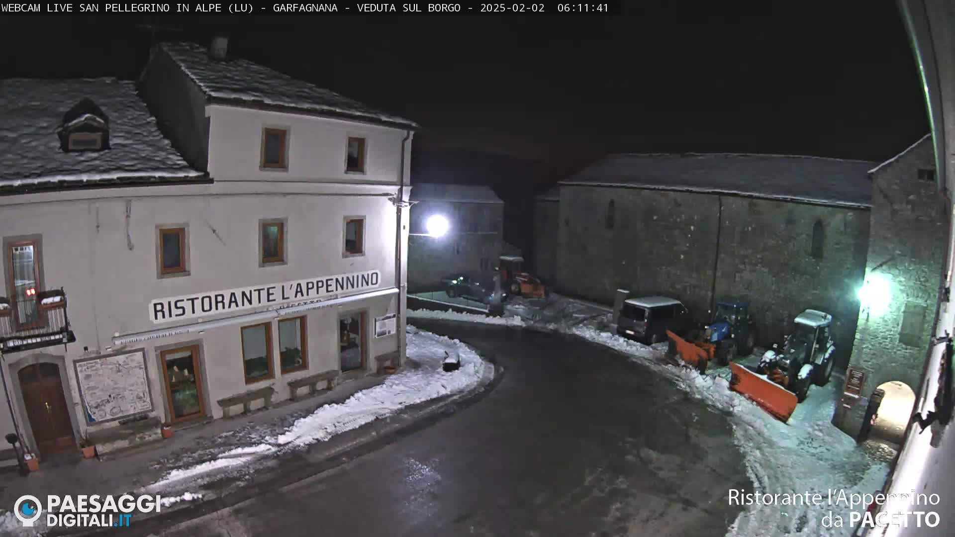 A snow-covered village square at night, illuminated by streetlights, features a restaurant and several snow-clearing vehicles.
