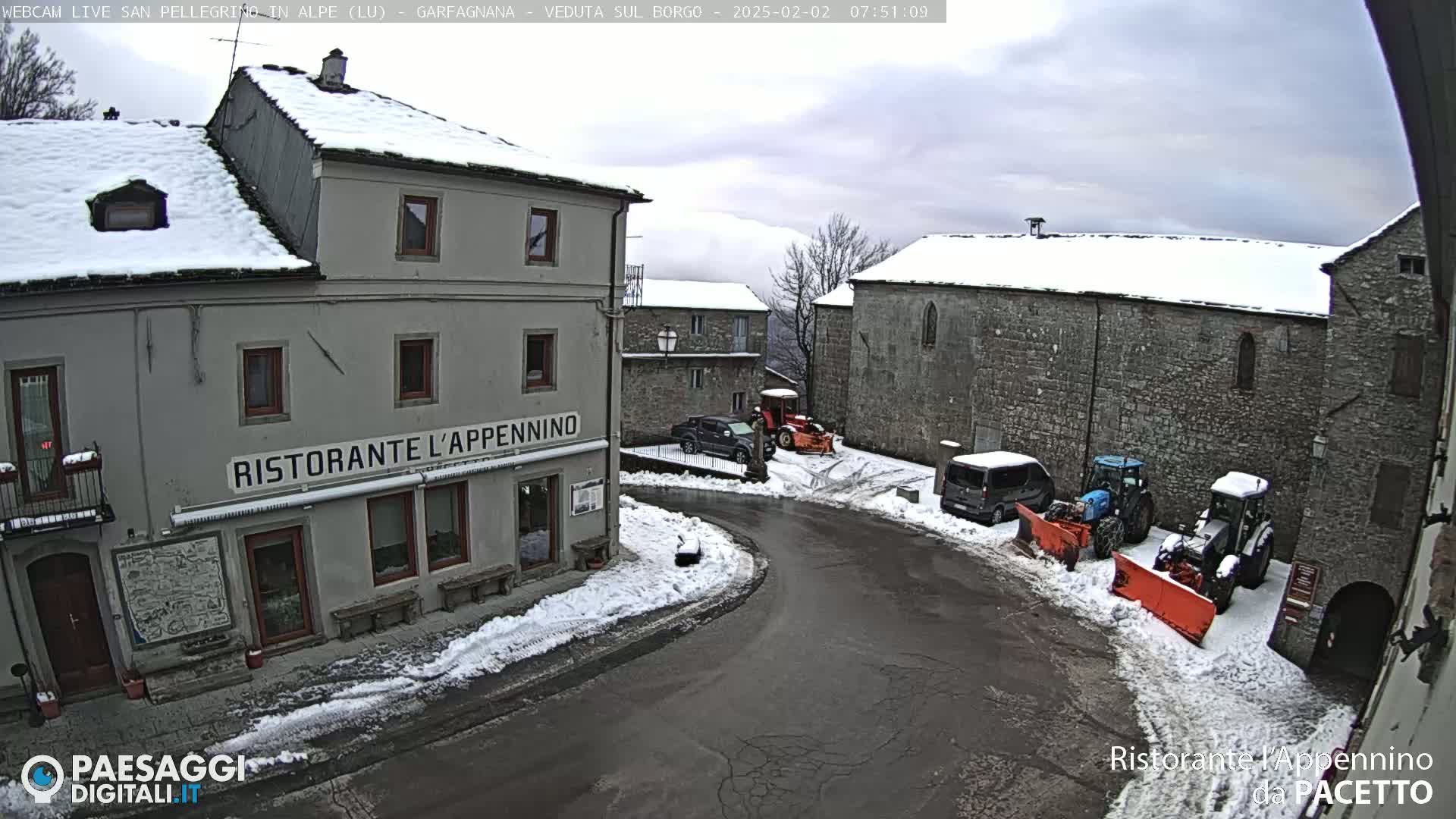 A snow-covered village square with stone buildings, parked vehicles, and snow removal equipment under a cloudy sky.