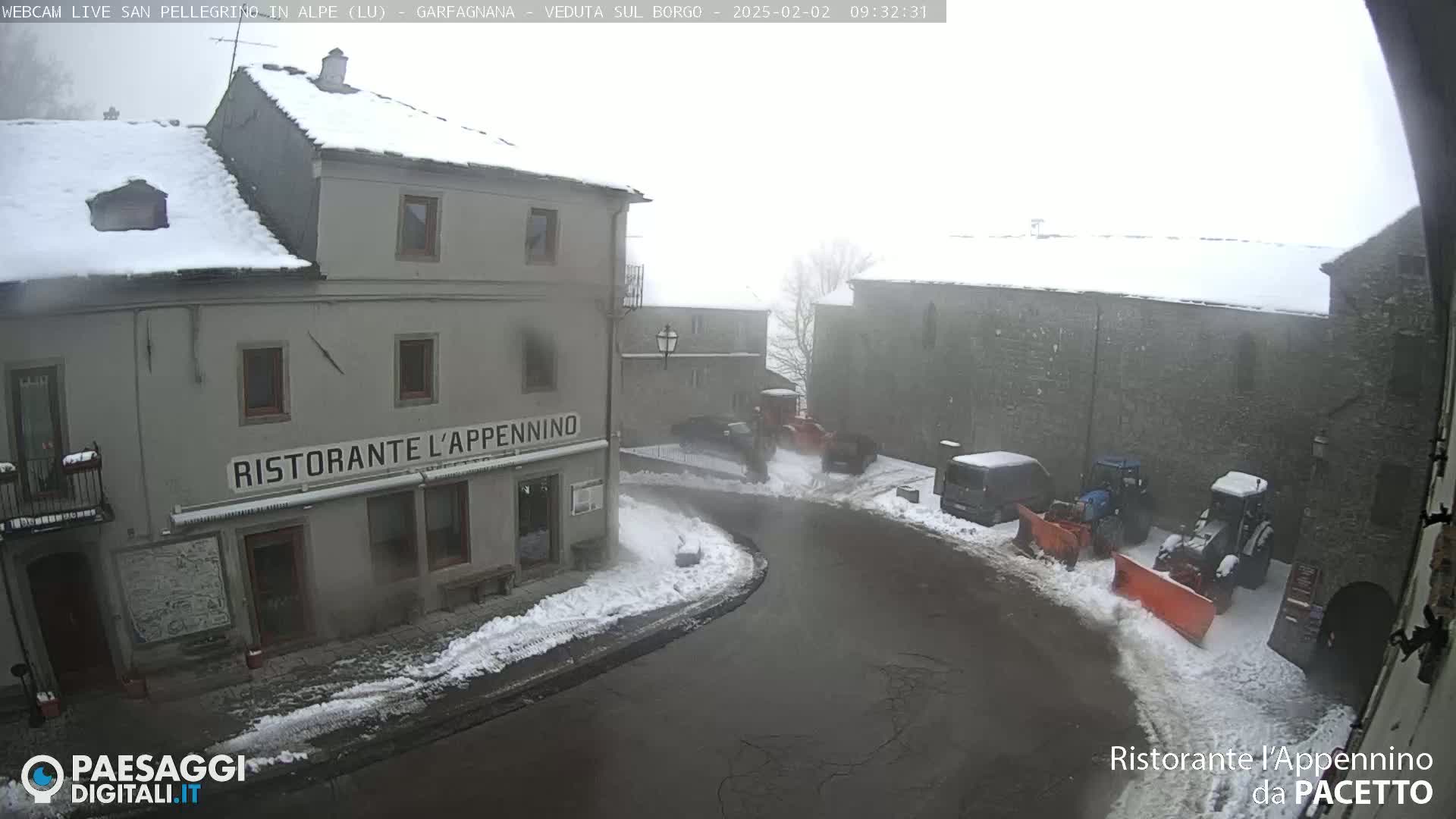 A snow-covered village street scene shows two buildings, several parked cars and snow removal equipment in foggy weather.