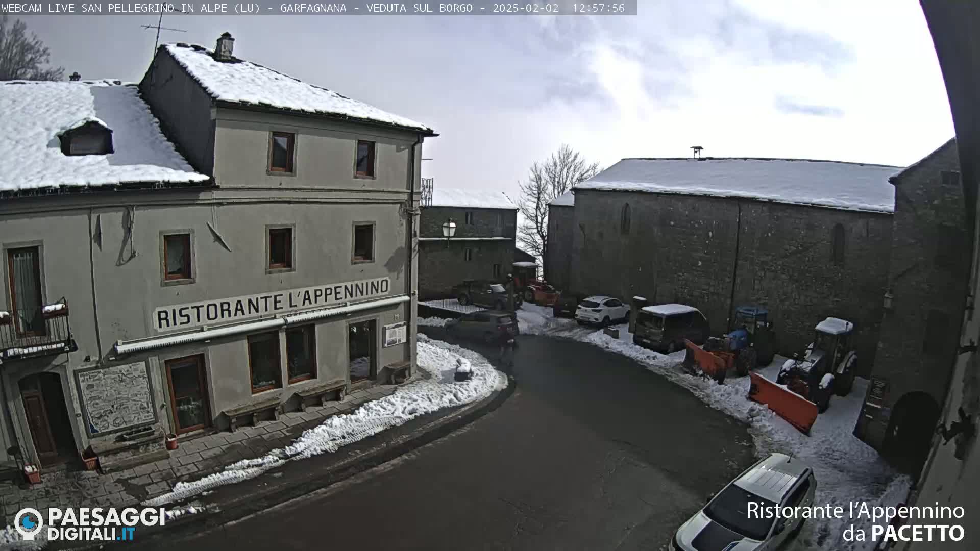 A snow-covered village square with cars parked along the sides, a restaurant, and some snow-clearing equipment.