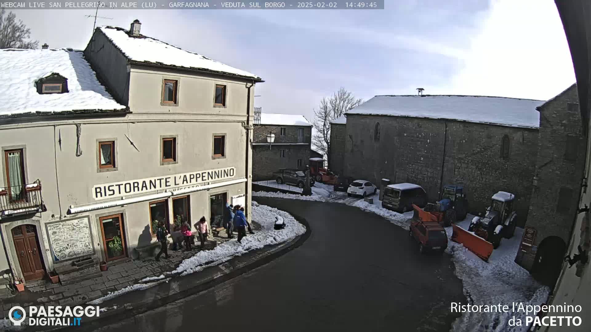 A sunny, snow-covered village square with several parked cars and some people near a restaurant.