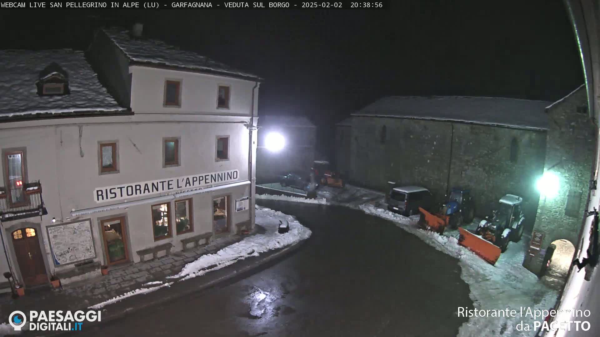 A snow-covered village square at night is illuminated, showing a restaurant, some parked vehicles, and snowplows clearing the street.