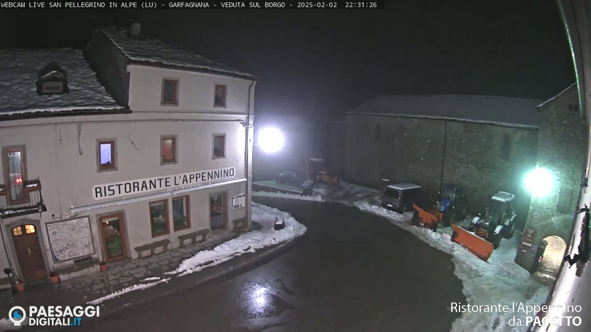 A snow-covered town square at night, with a restaurant, parked vehicles, and snow removal equipment.