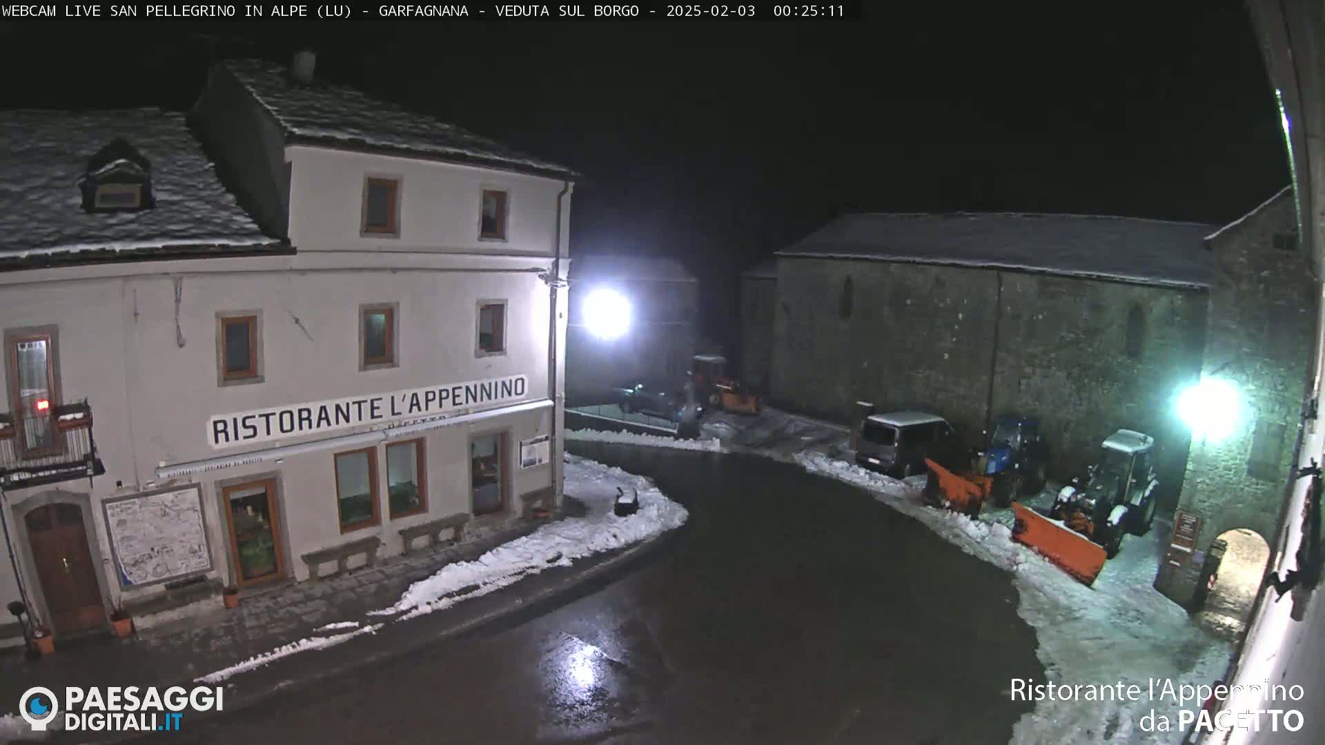 A snow-covered town square at night, illuminated by streetlights, shows a restaurant and several vehicles, including snow-removal equipment.