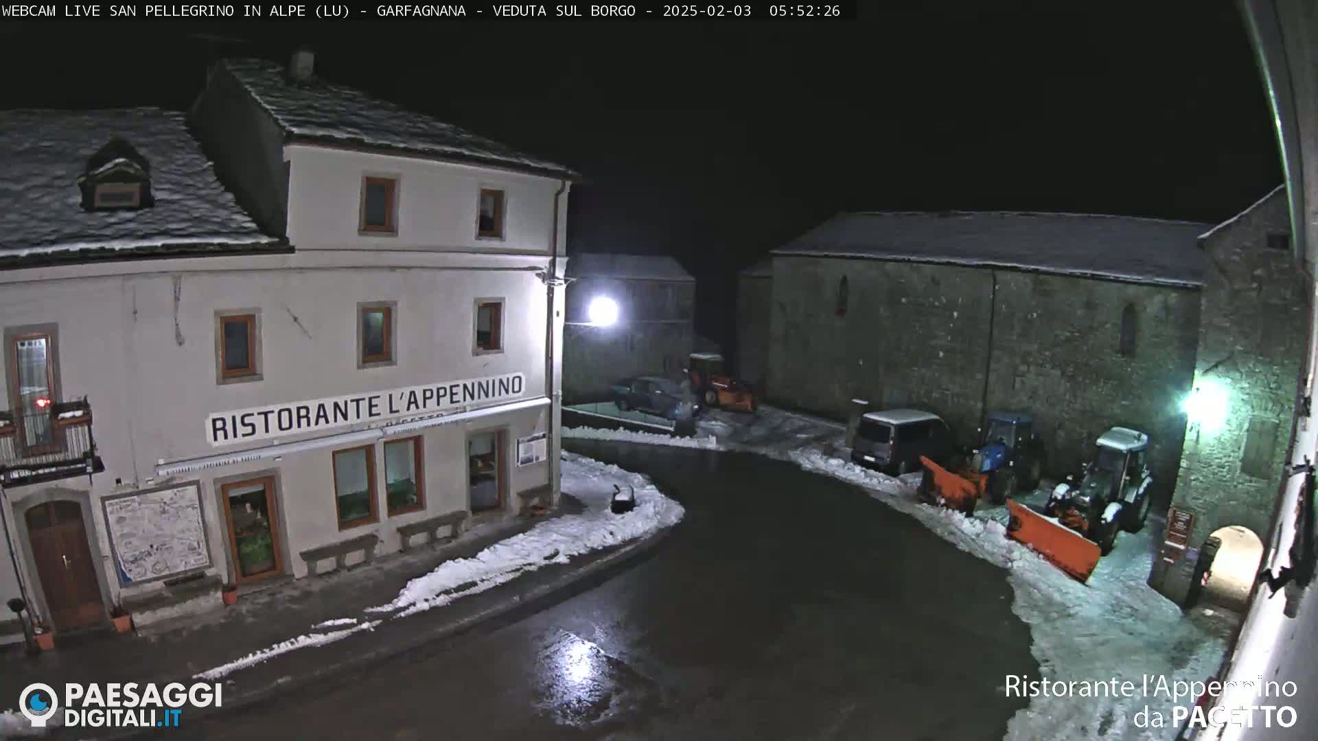 A snow-covered village square at night, illuminated by streetlights, shows several buildings, parked vehicles, and snow-clearing equipment.