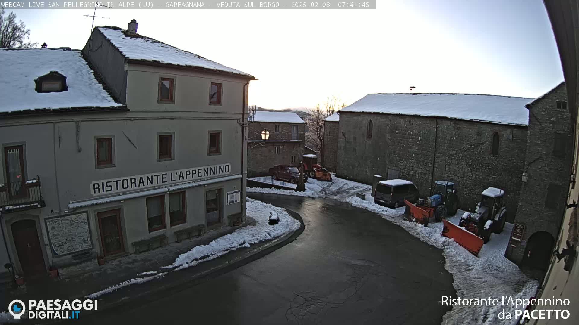 A snow-covered village square with several buildings, some vehicles, and snow-clearing equipment, under a clear, bright sky.