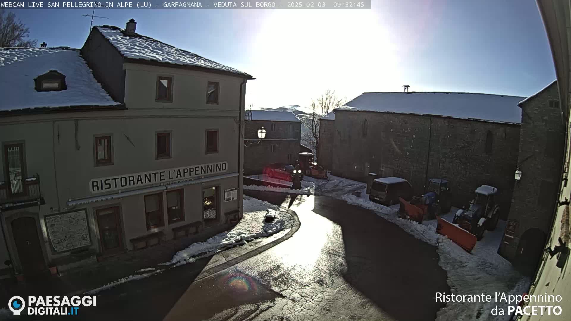 A snow-covered village square with several buildings, a restaurant, and some parked vehicles under a bright, sunny sky.