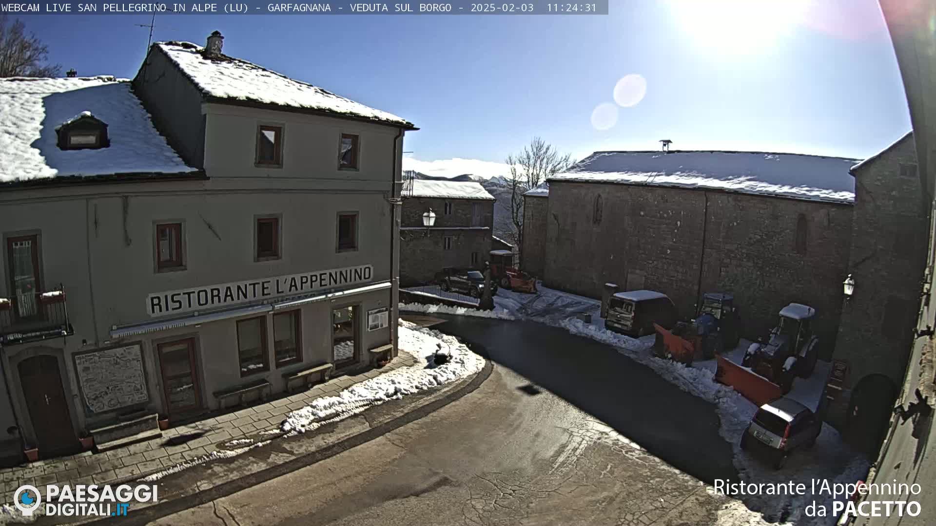 A snow-covered village square with several buildings, some vehicles, and snow plows under a sunny sky.