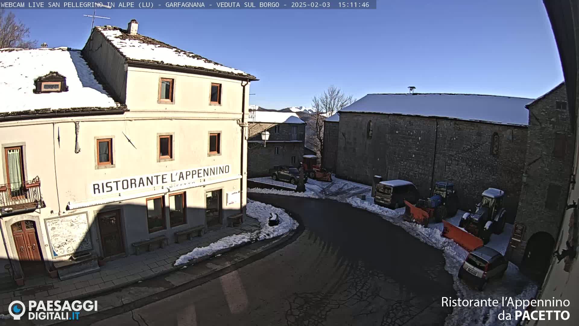 A sunny winter scene shows a snow-covered village square with several parked vehicles and snow removal equipment, including tractors with snow plows.