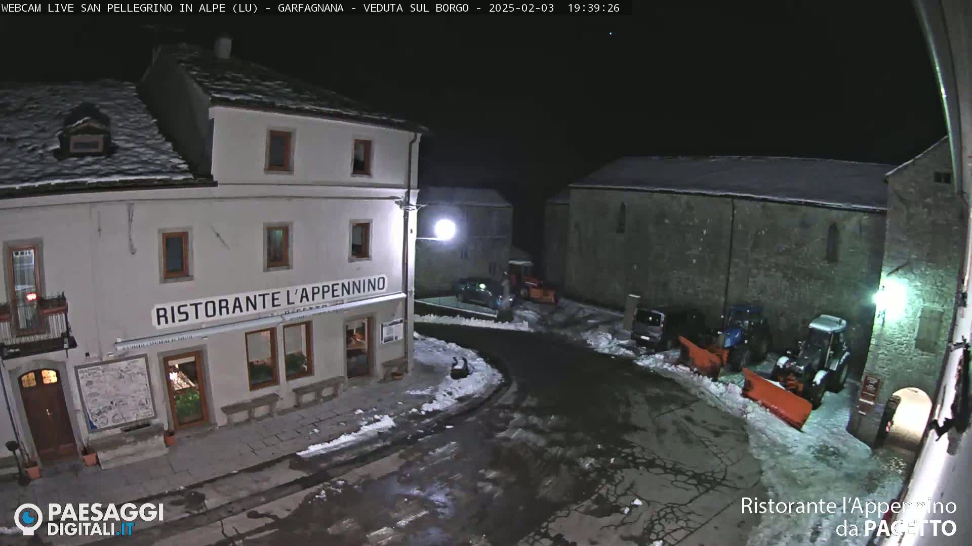 A nighttime snow-covered town square scene shows a restaurant, some parked vehicles, and snow removal equipment.