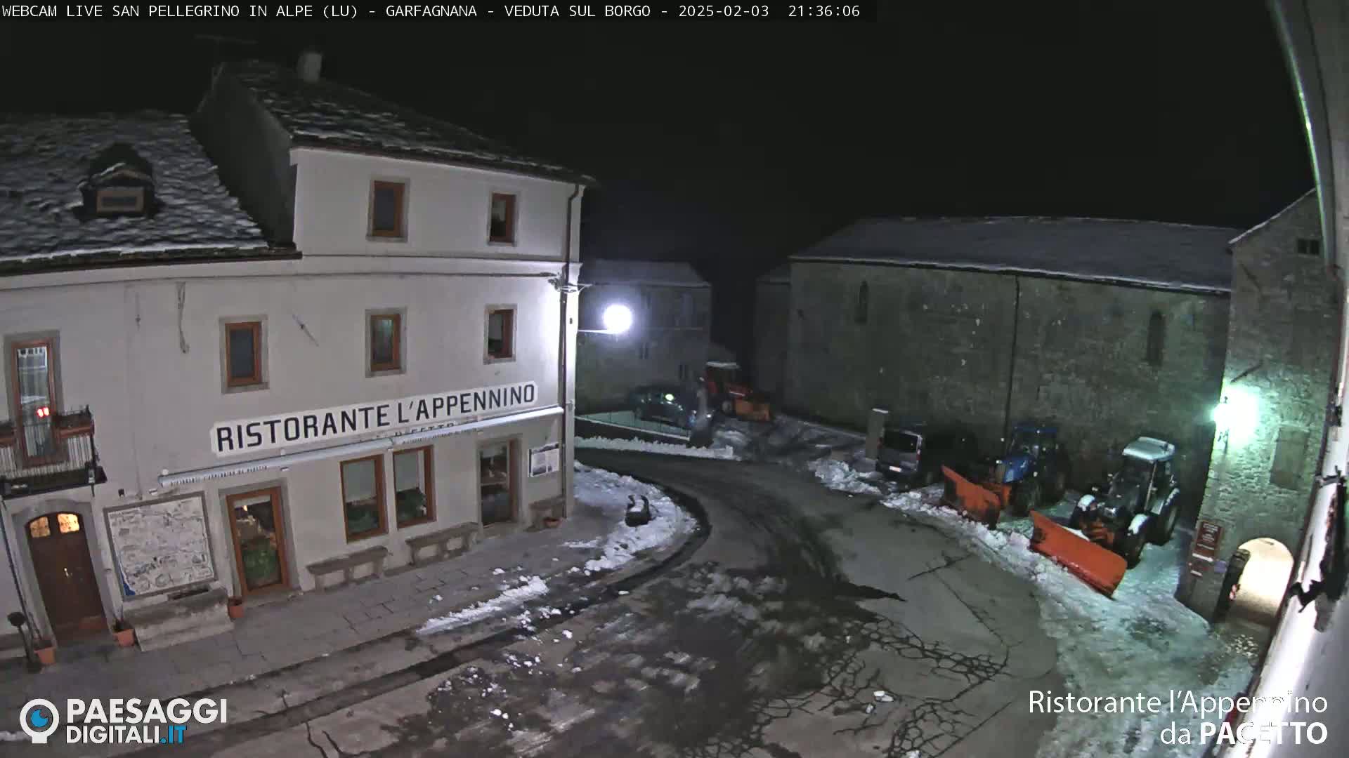 A snow-covered village square at night, illuminated by streetlights, shows a restaurant and several parked vehicles, with snowplows nearby.