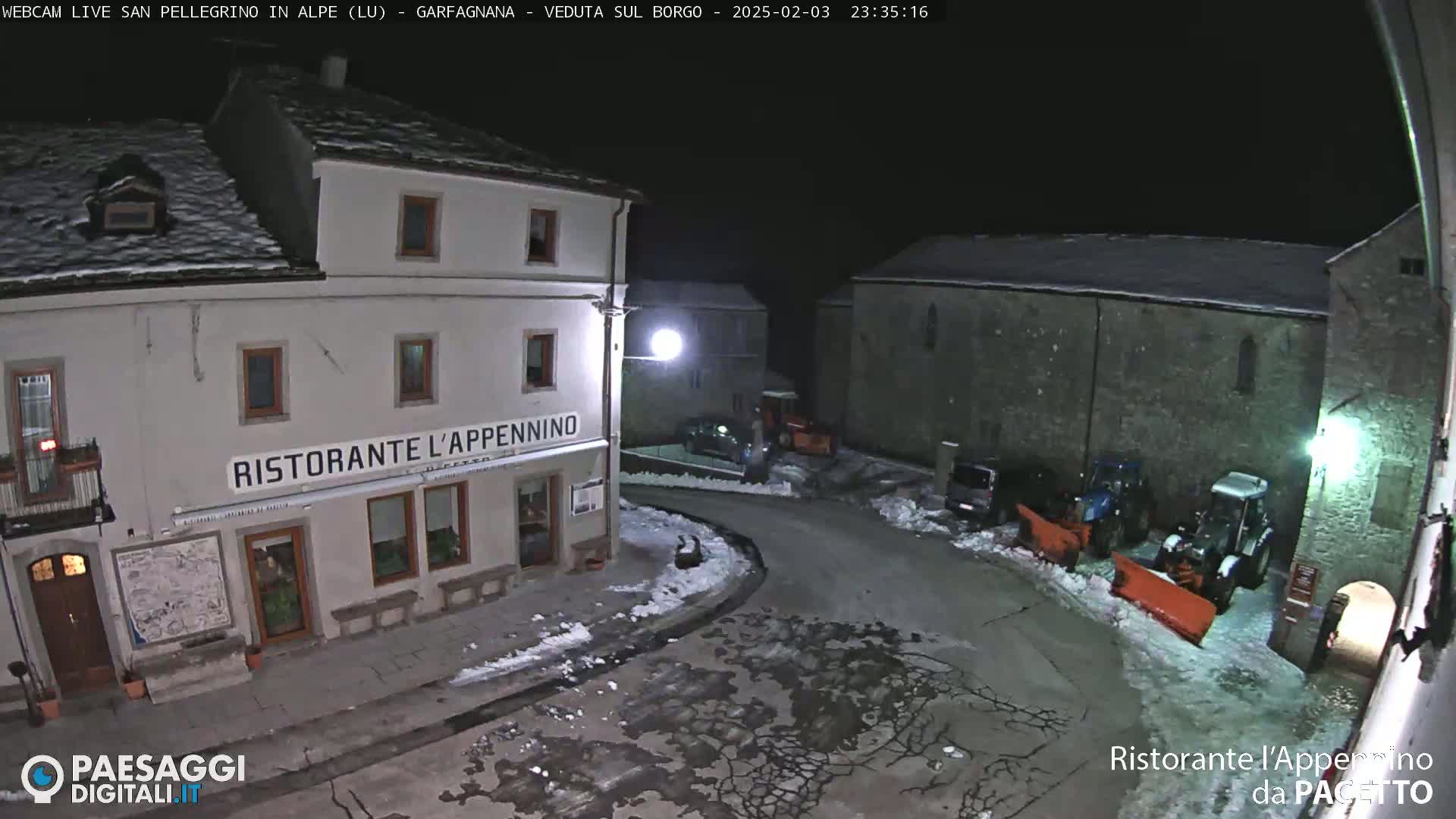 A snow-covered village square at night, illuminated by a single lamppost, shows a restaurant and several parked vehicles with snow plows nearby.